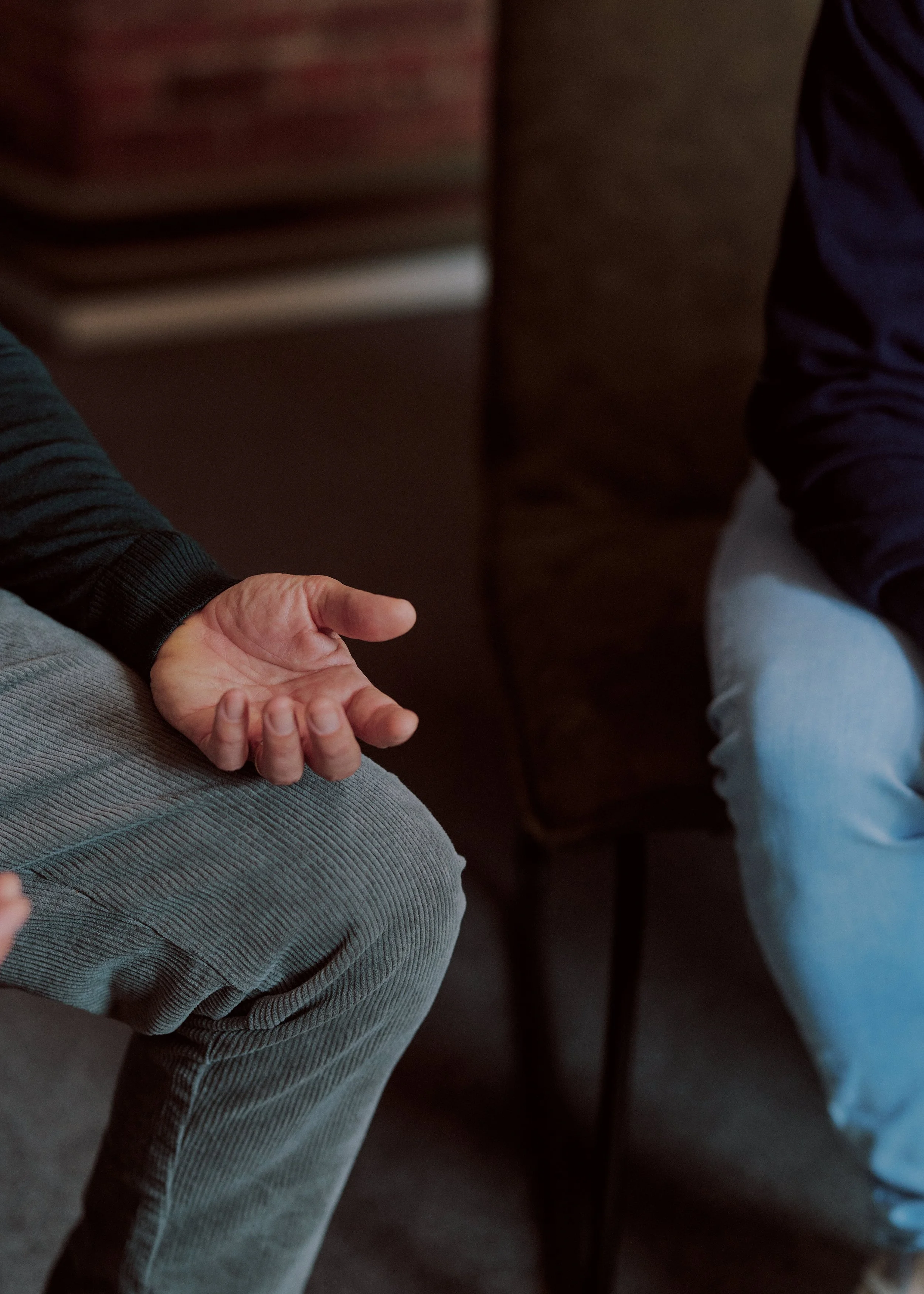 Close-up of one person's hand resting on their lap, with two other people around, one on the left and one on the right, in a dimly lit setting.