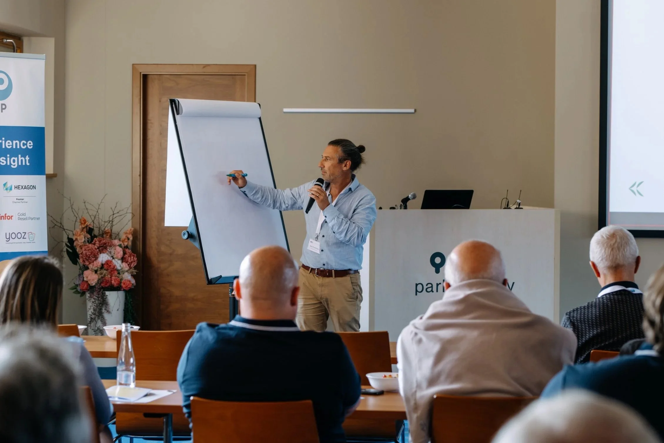 A man with long hair tied back, wearing a light blue shirt, is giving a presentation at a conference. He is holding a microphone in one hand and a blue marker in the other, writing on a flip chart. There are several audience members seated at tables, some with water bottles and bowls. A large banner and a podium with the logo 'par...' and some flowers are visible in the background.