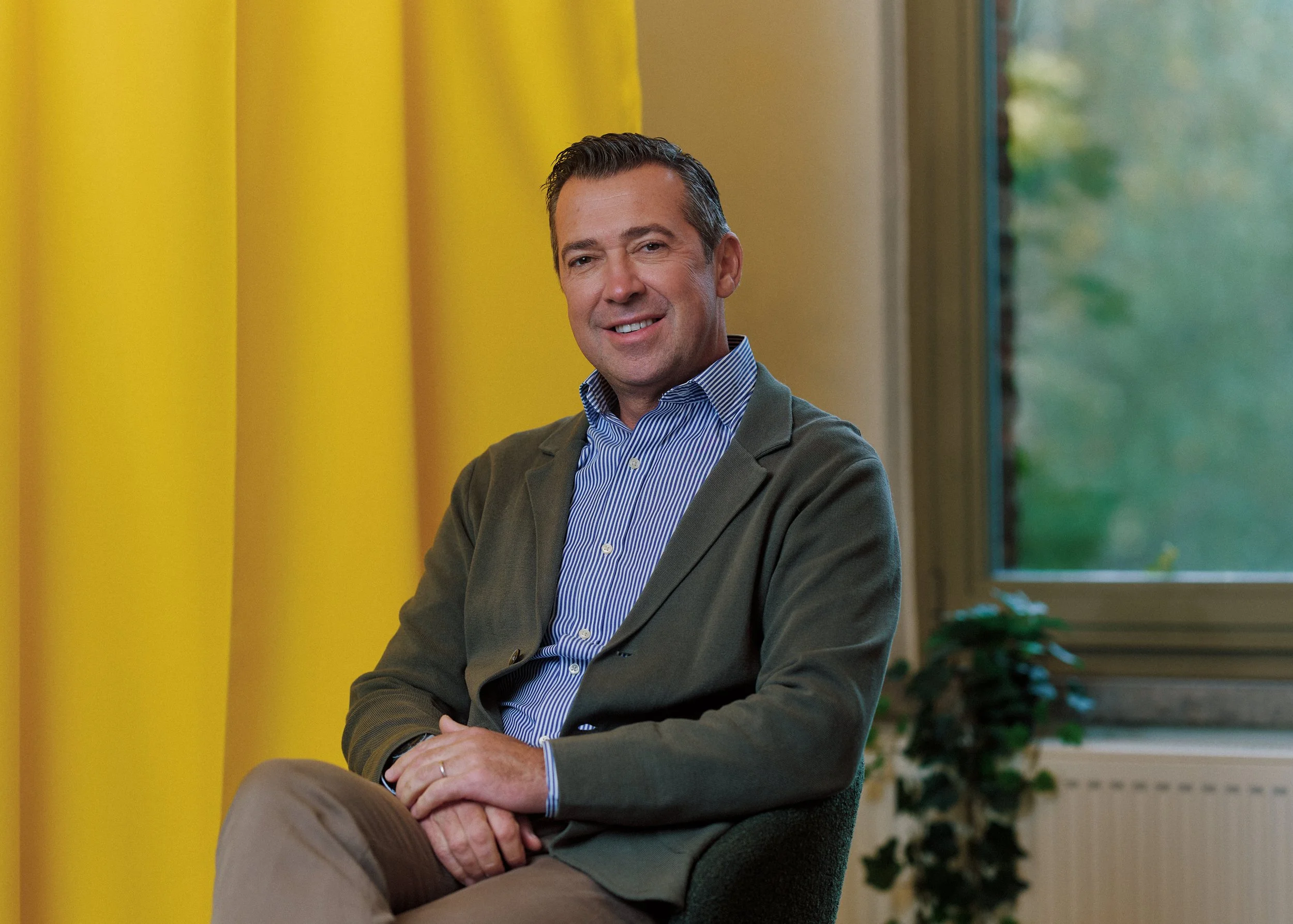 A middle-aged man with dark hair wears a gray blazer over a striped shirt. He smiles and sits with one leg crossed, hands resting on his knee, in front of a yellow curtain and a window with green trees outside.