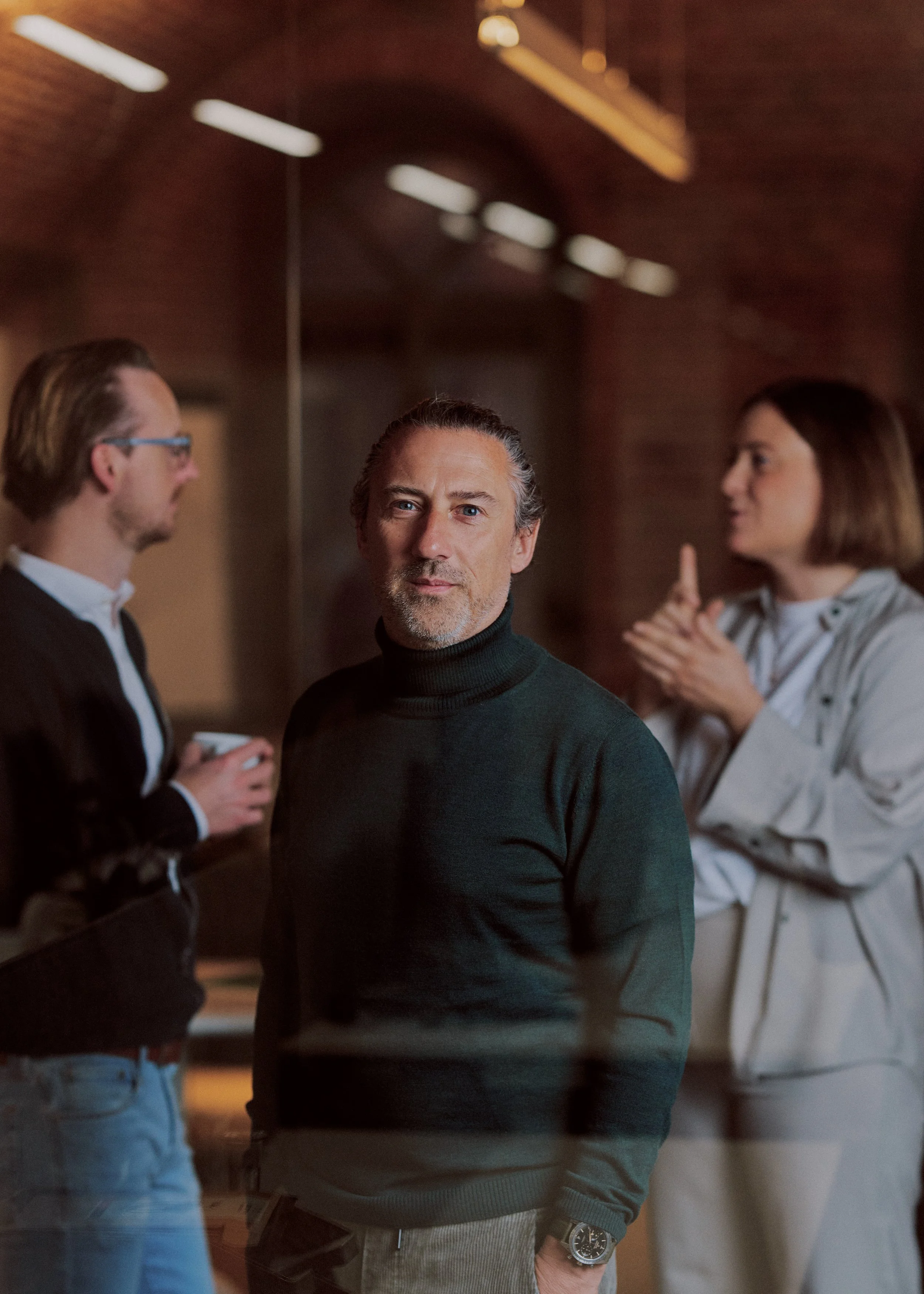 A middle-aged man with gray hair and a beard, wearing a dark turtleneck sweater and a wristwatch, stands in a dimly lit room. He is surrounded by two women and another man, who are engaged in conversation.