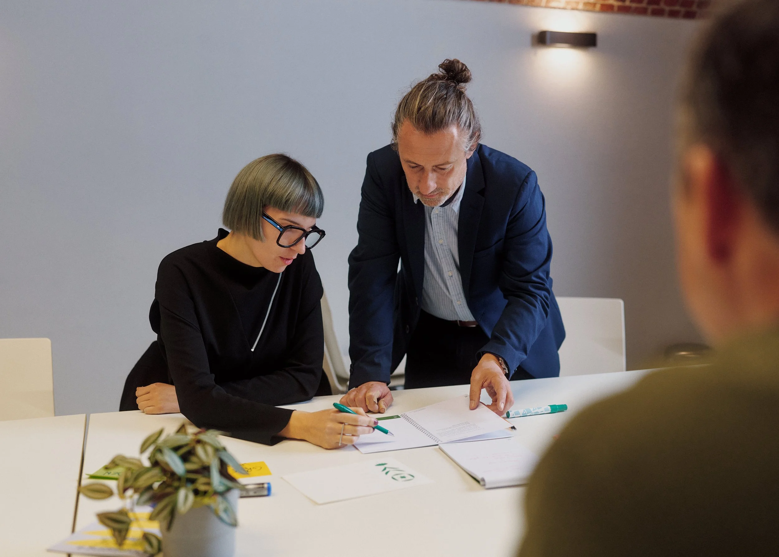 Two people, a woman with glasses and a man with a bun hairstyle, are engaging in a discussion over documents at a white table. The woman is seated, writing in a notebook, while the man leans over, pointing at something on the document. The setting appears to be an office or meeting room with a gray wall and soft lighting.