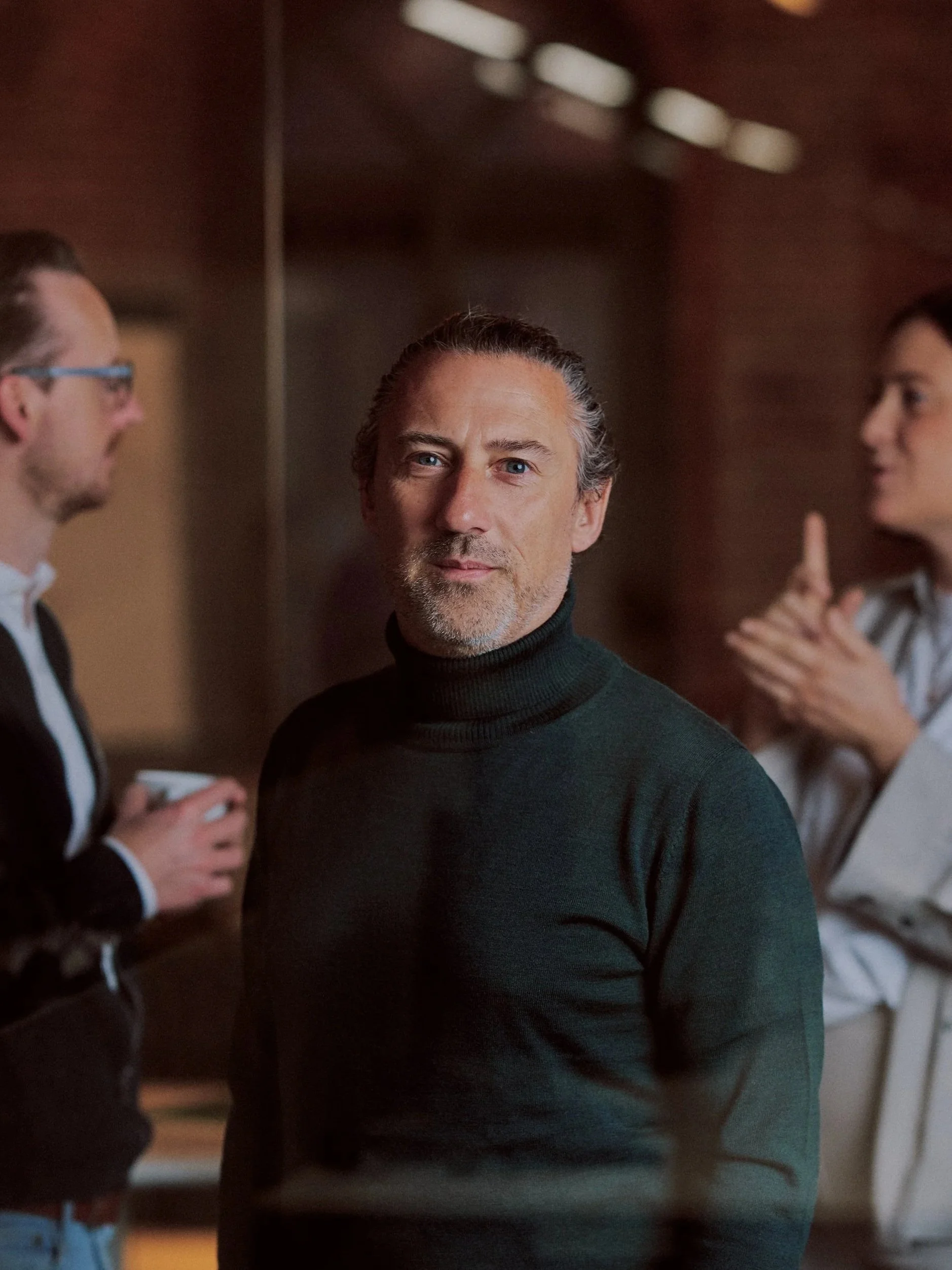 A middle-aged man with gray hair and a beard, wearing a dark turtleneck, standing in front of a mirror with two women behind him, one of whom is wearing glasses and talking, and the other is gesturing with her hands.