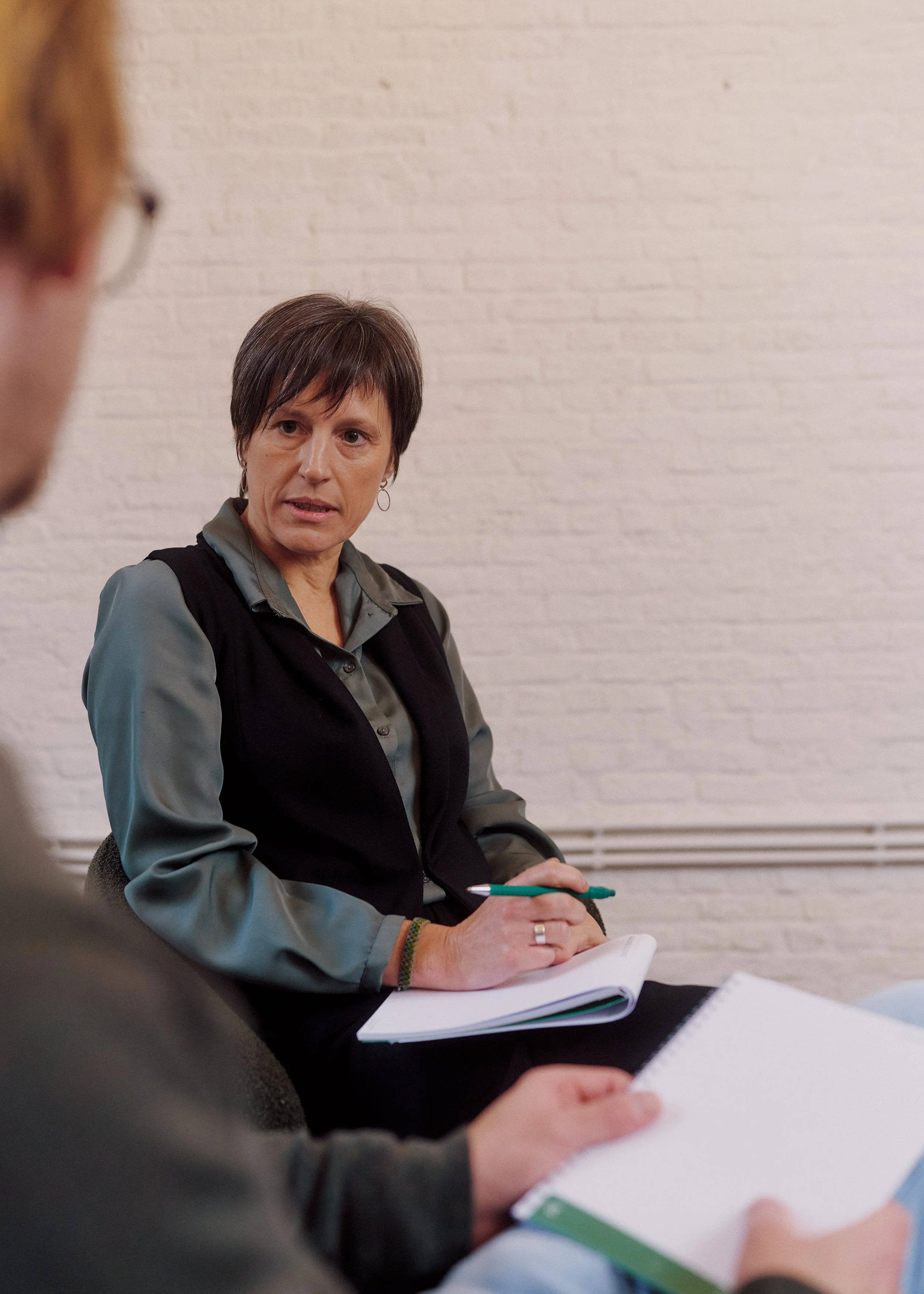 A woman with short dark hair, wearing a gray blouse and black vest, sitting at a table during a meeting, holding a green pen and taking notes, with a white brick wall in the background.