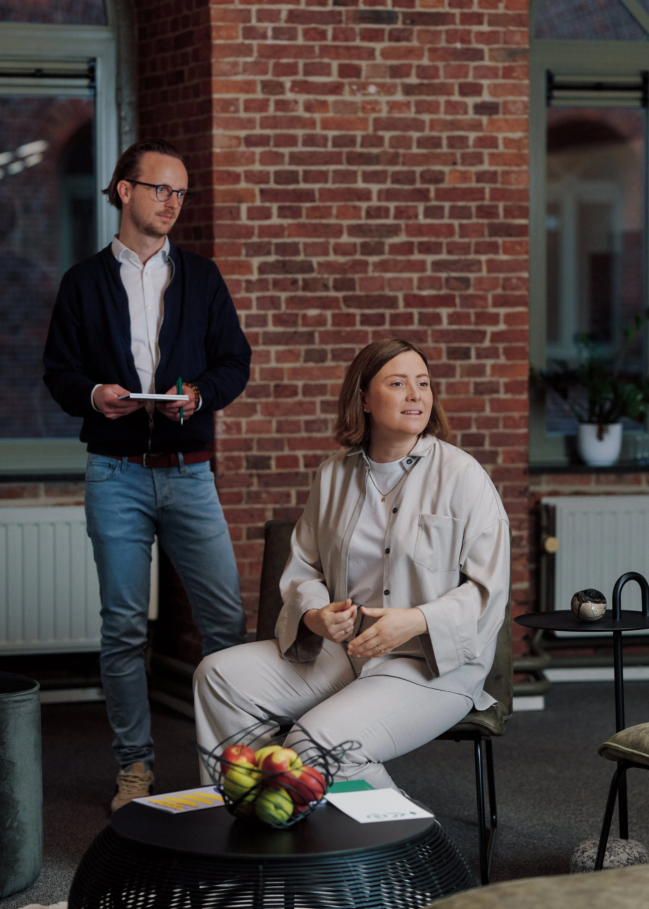 A man with glasses, wearing a white shirt and black jacket, stands with a notebook and pen in his hands. A woman with shoulder-length brown hair, wearing a beige outfit, is sitting on a chair, looking to the side. They are in a room with brick walls, large windows, and indoor plants, possibly a casual meeting or workspace.