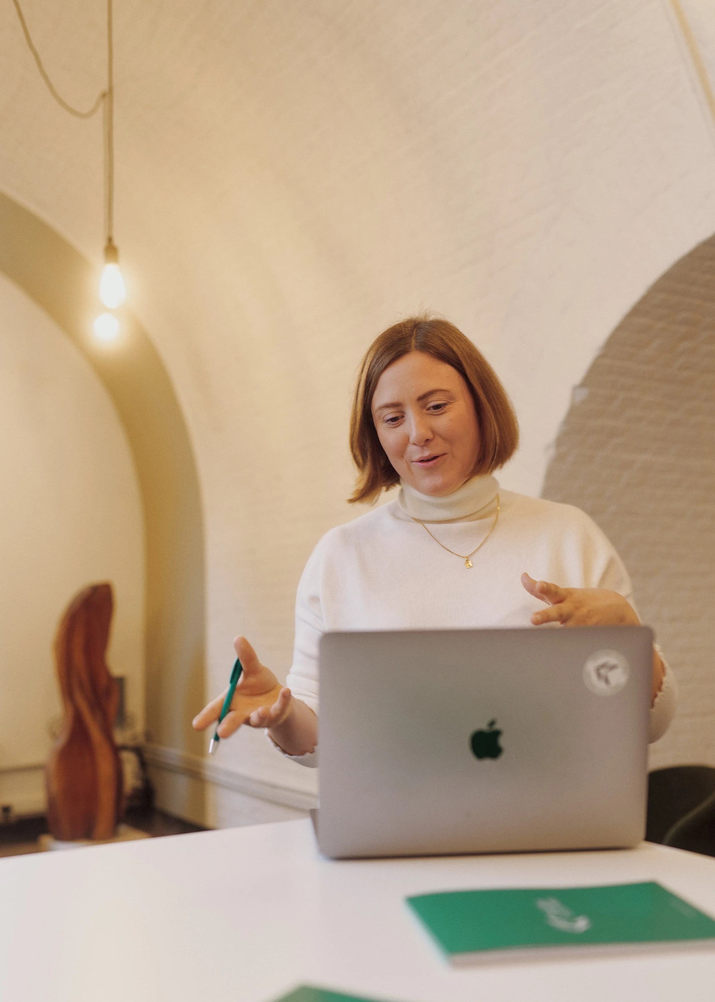 A woman with shoulder-length brown hair, wearing a cream turtleneck sweater and a gold necklace with a small pendant, sitting at a table with a silver laptop. She is smiling and gesturing with her hands, possibly engaged in a conversation or presentation, with a blurred background of a cream-colored wall and warm lighting.
