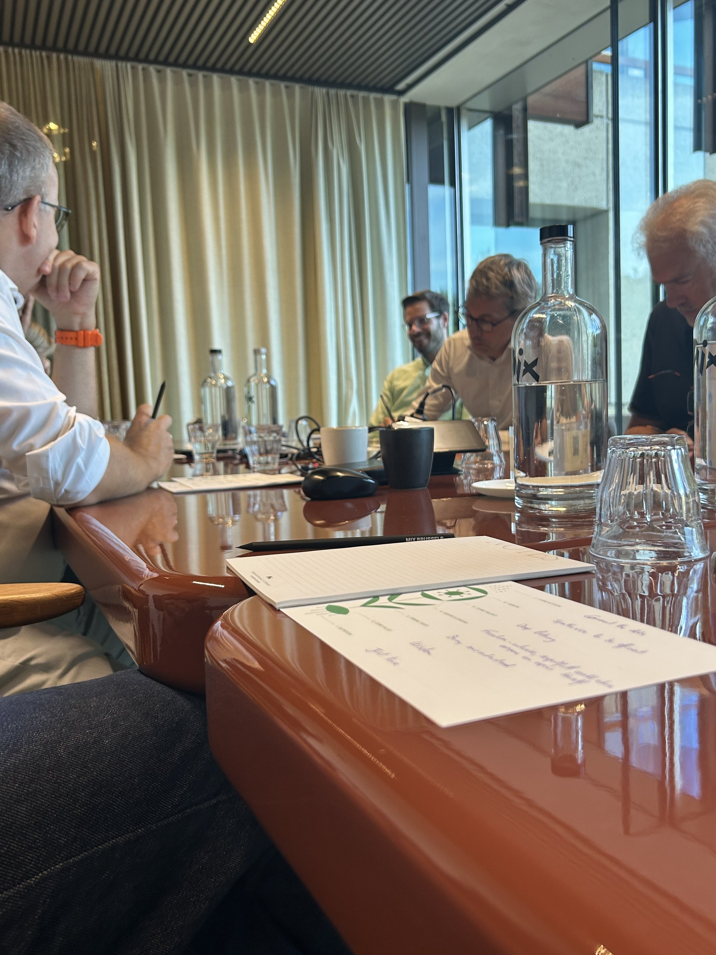 People sitting around a conference table with documents, water bottles, and glasses, in a modern room with large windows and beige curtains.