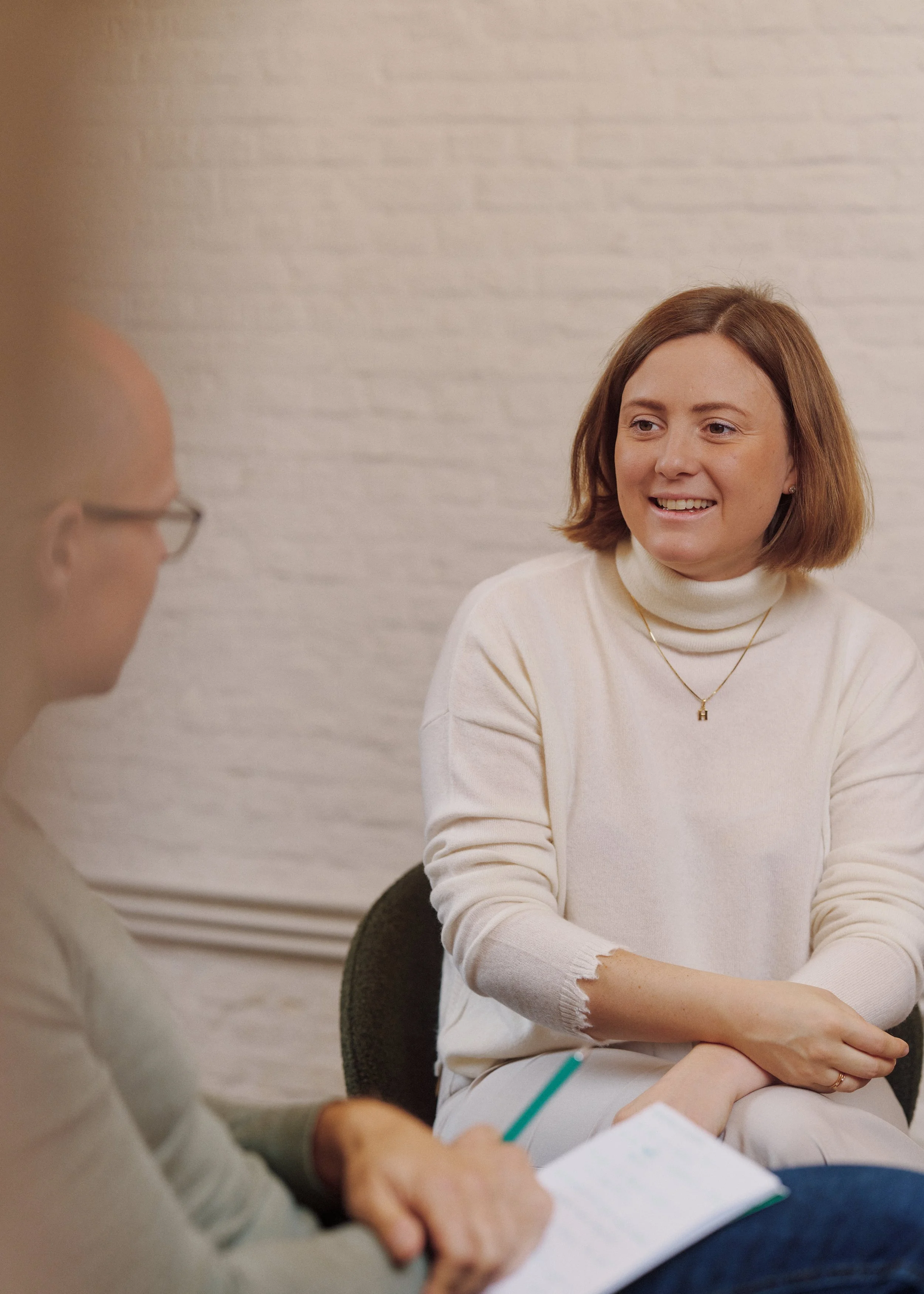 Two women sitting at a table engaged in a conversation, with one woman smiling and listening, wearing a cream turtleneck sweater and a gold necklace with an 'H' pendant.
