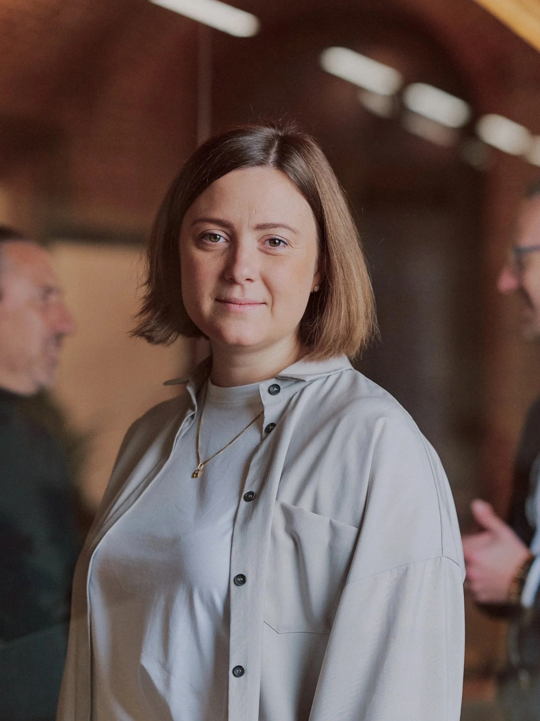 A young woman with short brown hair, wearing a light grey button-up shirt and a gold necklace, standing and looking directly at the camera with a slight smile. Blurred people are visible in the background.