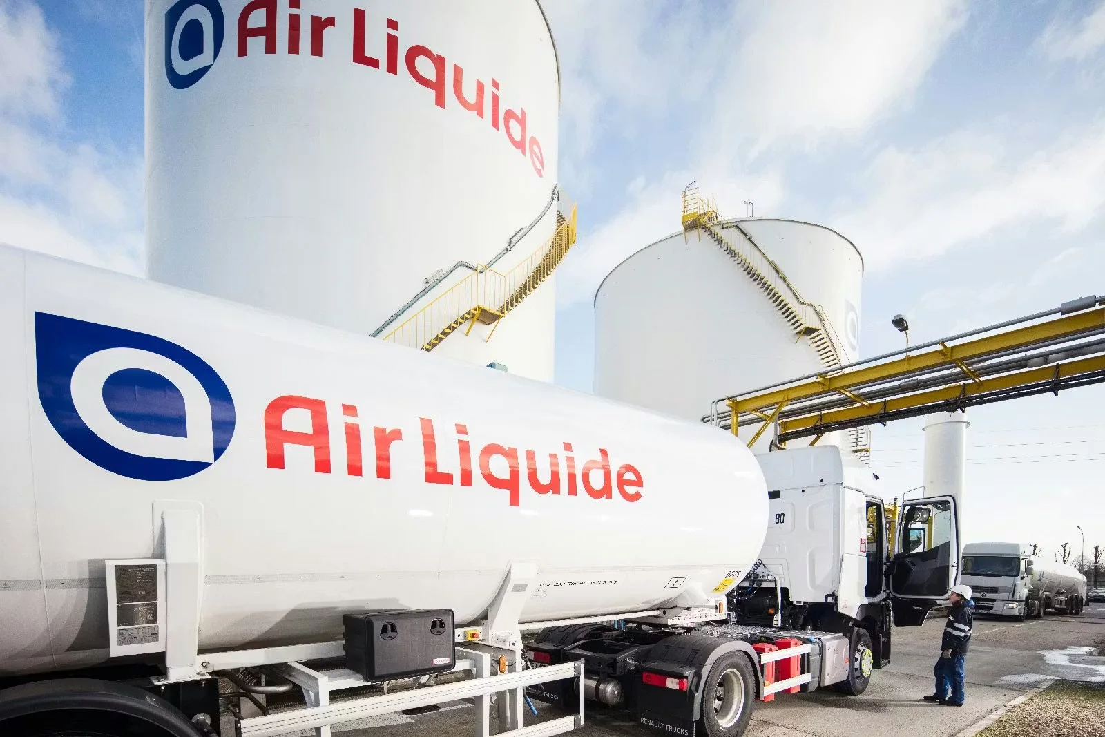 Cannery trucks with the Air Liquide logo parked outside with large storage tanks in the background under a partly cloudy sky.