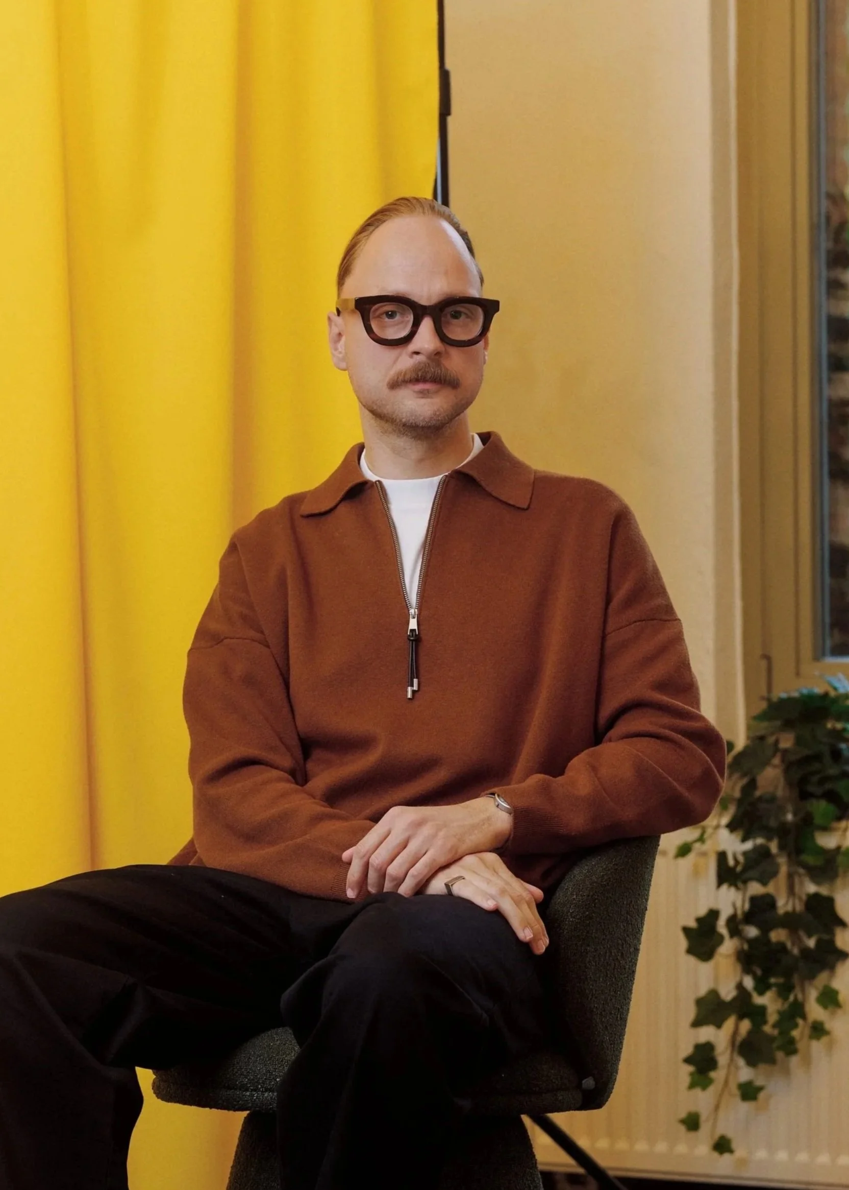 A man with red hair and glasses sitting in front of a yellow curtain and plant, wearing a brown jacket and black pants.