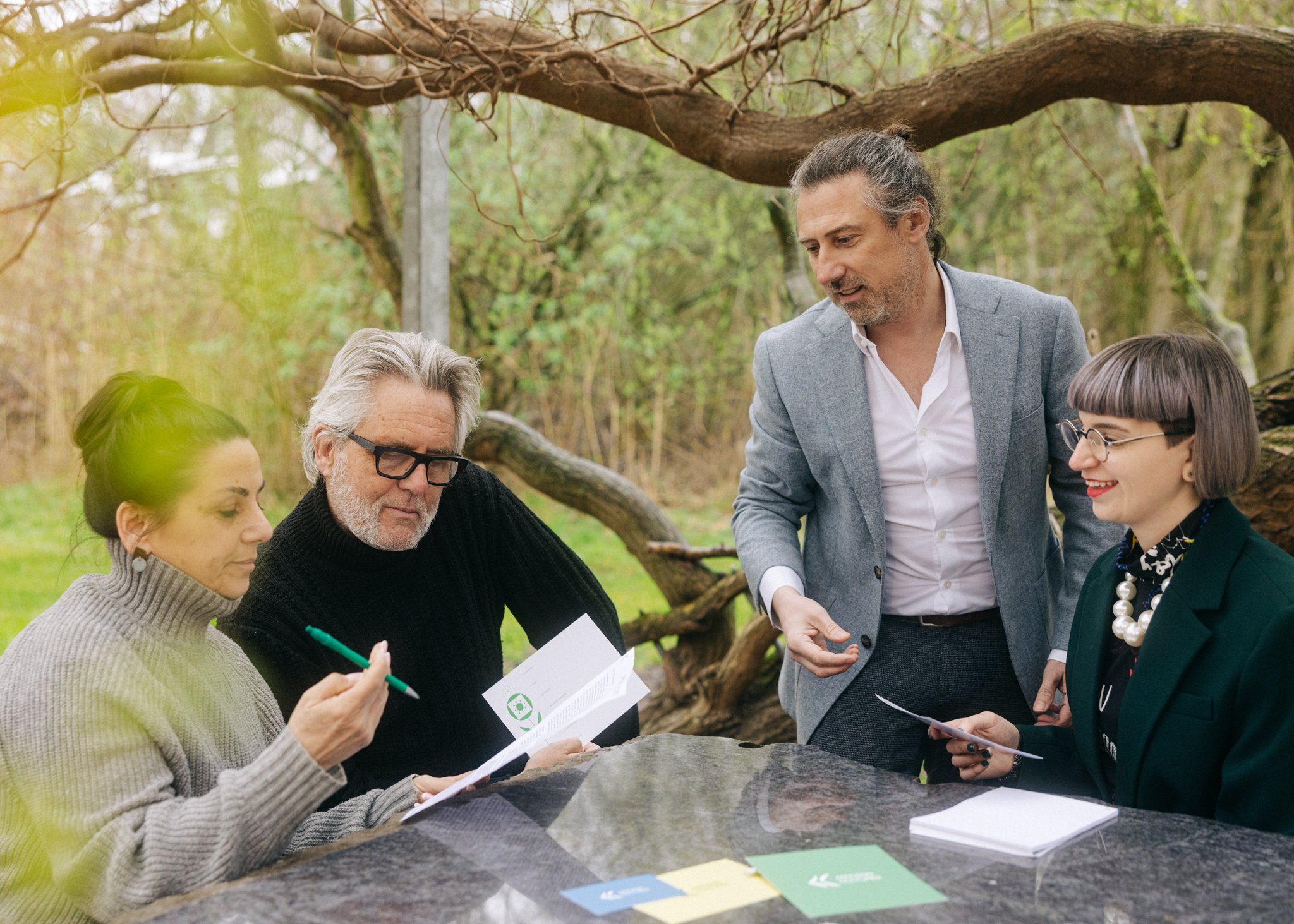 Group of five people engaged in a discussion outdoors in a forested area, looking at papers and charts on a large flat rock.