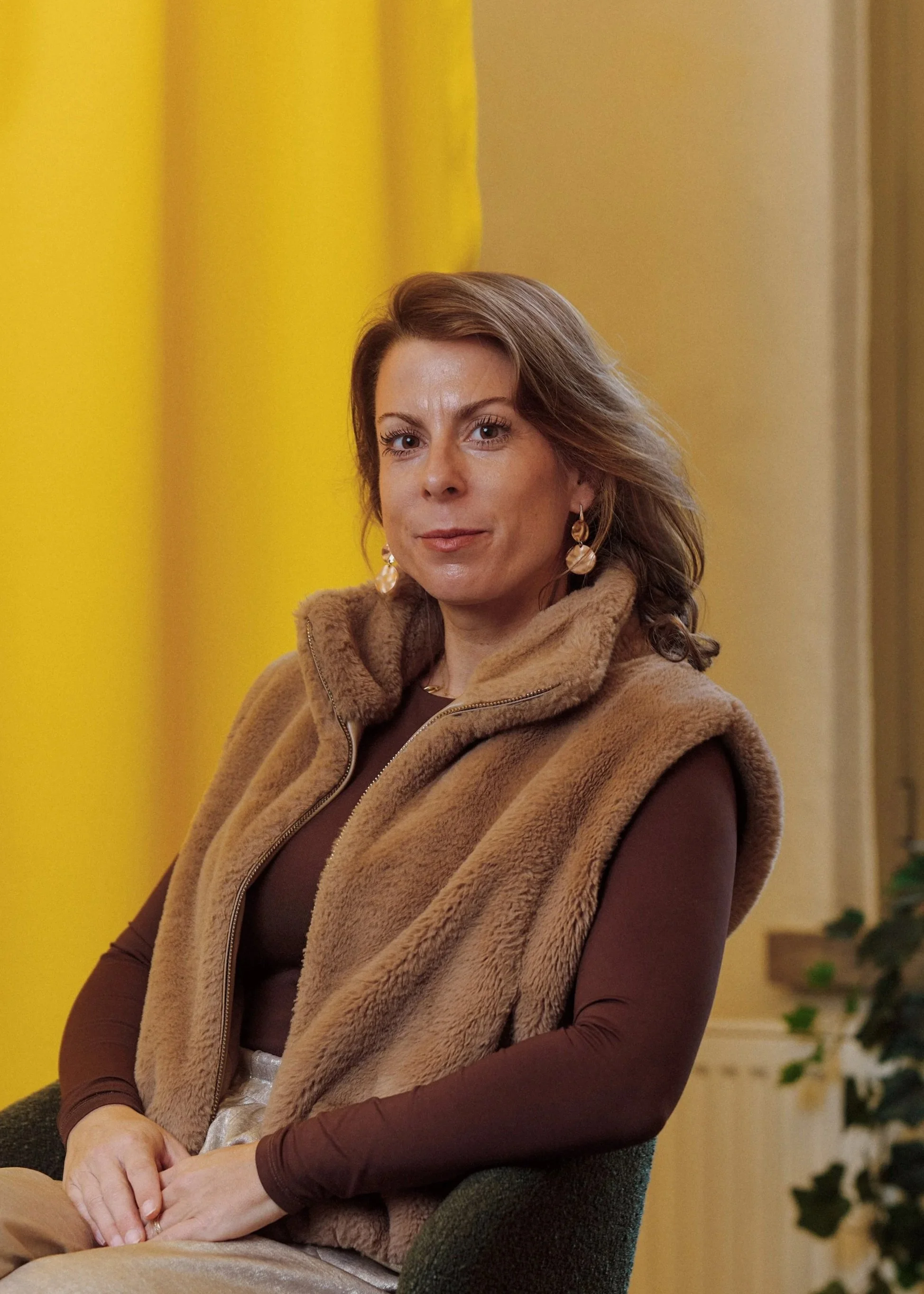 A woman sitting on a gray chair wearing a brown fleece vest over a long sleeve brown shirt, with earrings, in a room with yellow and beige curtains.