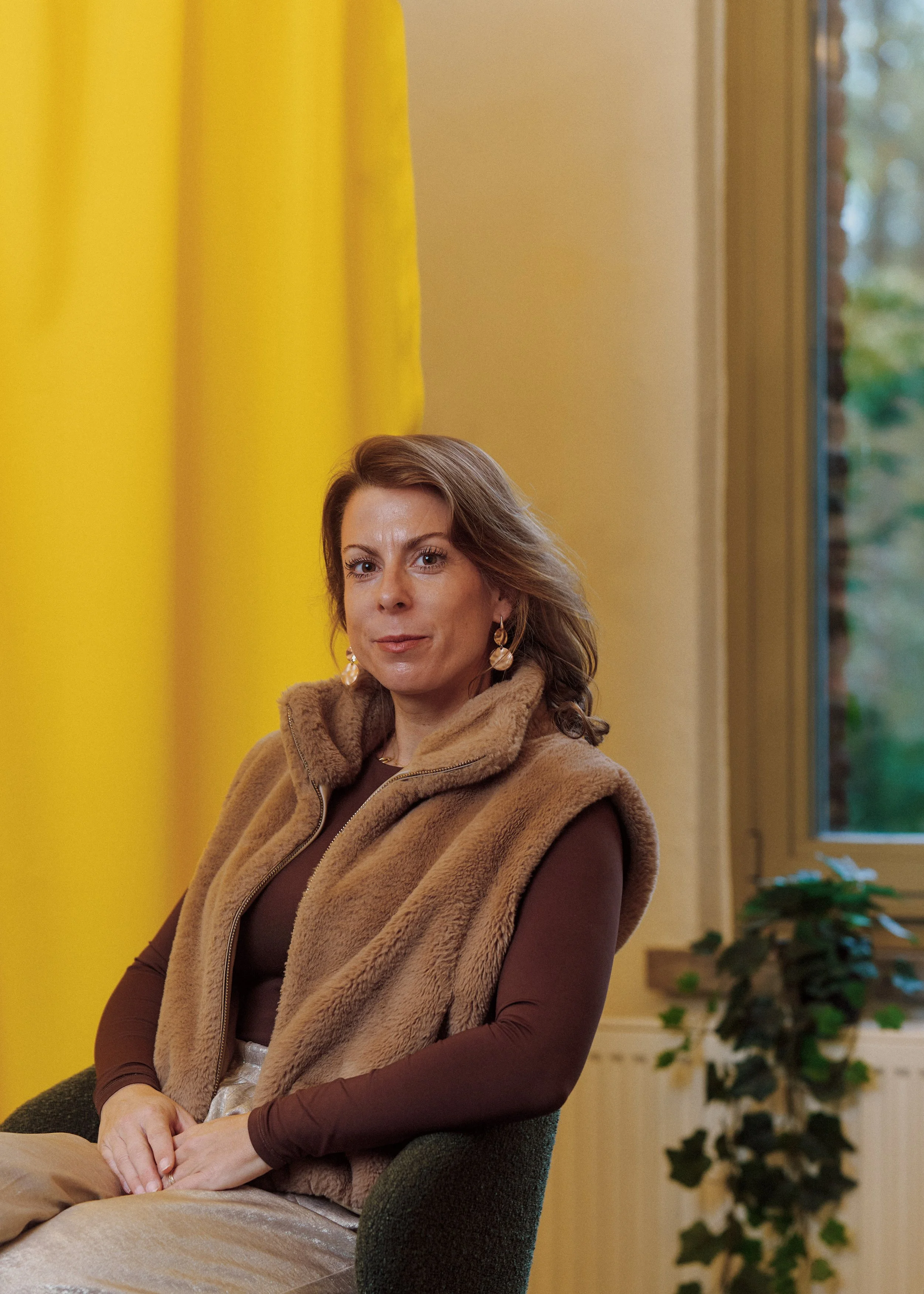 A woman sitting in a room with yellow walls, a window, and houseplants, wearing a brown long-sleeve top with a fur vest and earrings.