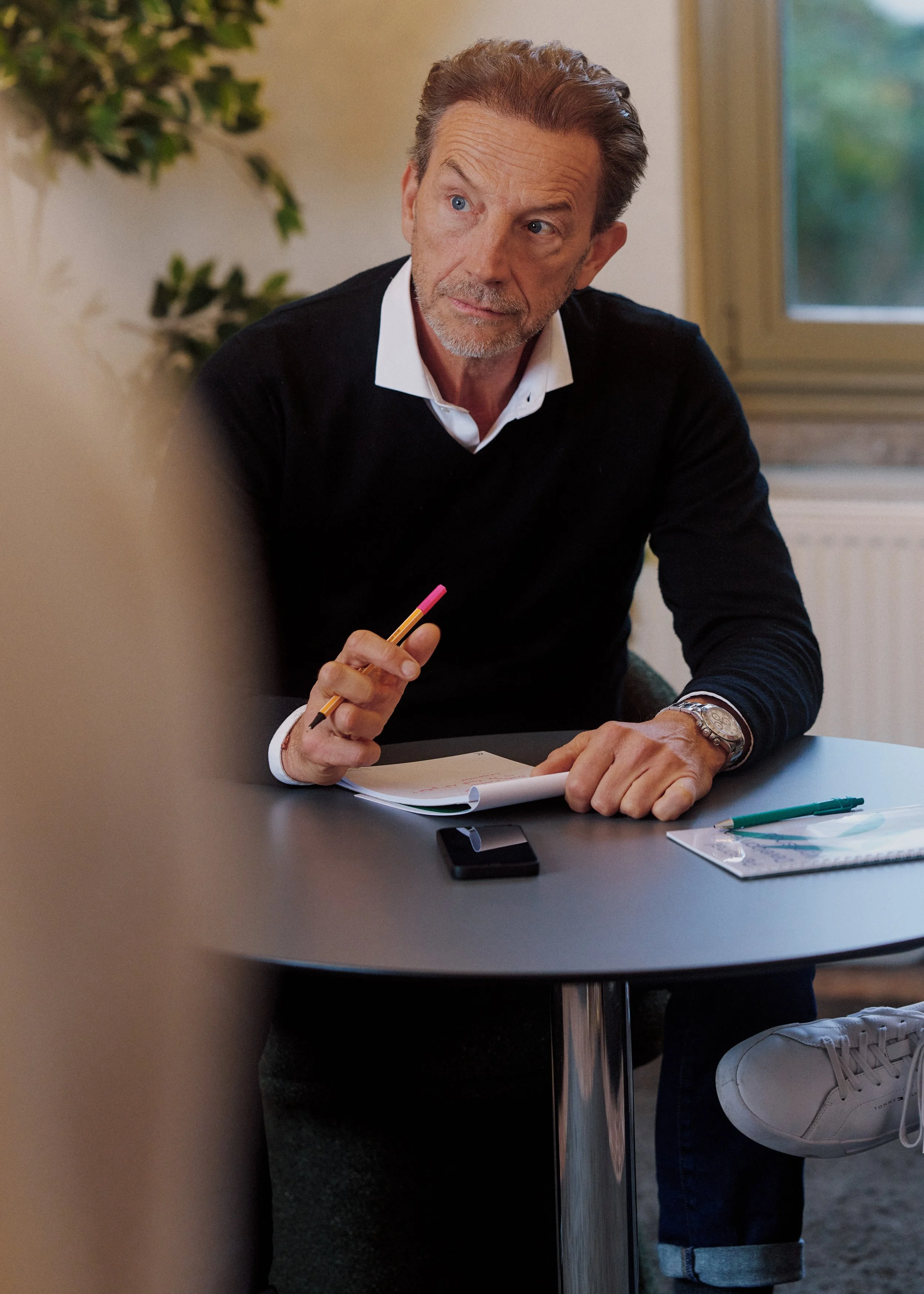 A man with brown hair and a beard wearing a black sweater and white shirt, sitting at a table with papers, a pen, and a smartphone, holding a pencil while talking.