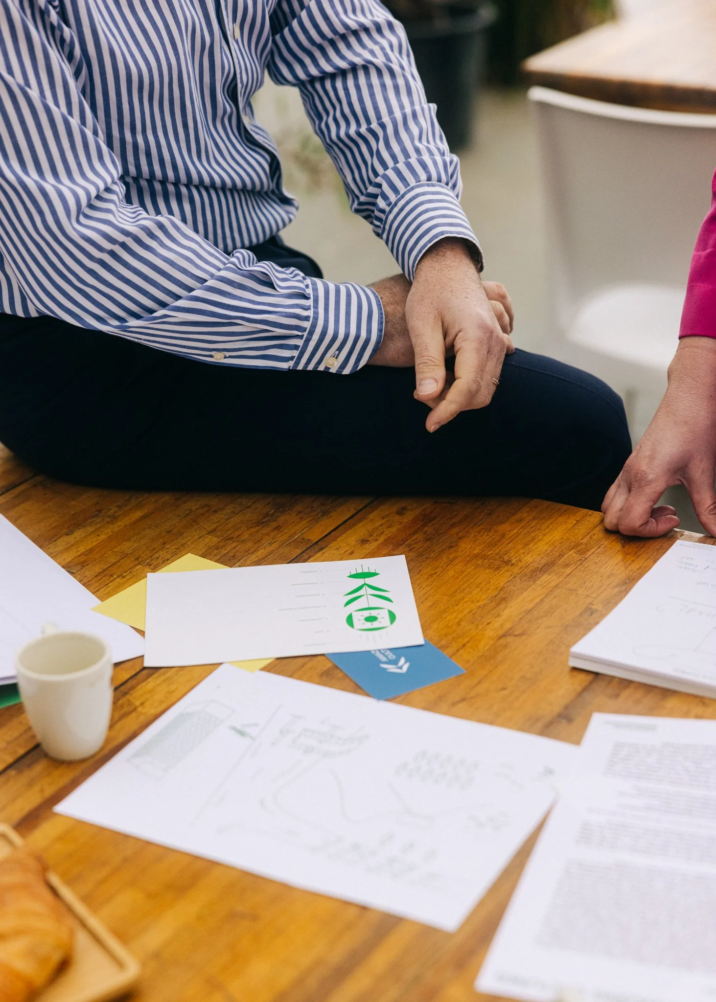 A person wearing a striped shirt is sitting cross-legged at a wooden table, with various papers and diagrams spread out, and a small paper cup is visible on the table.