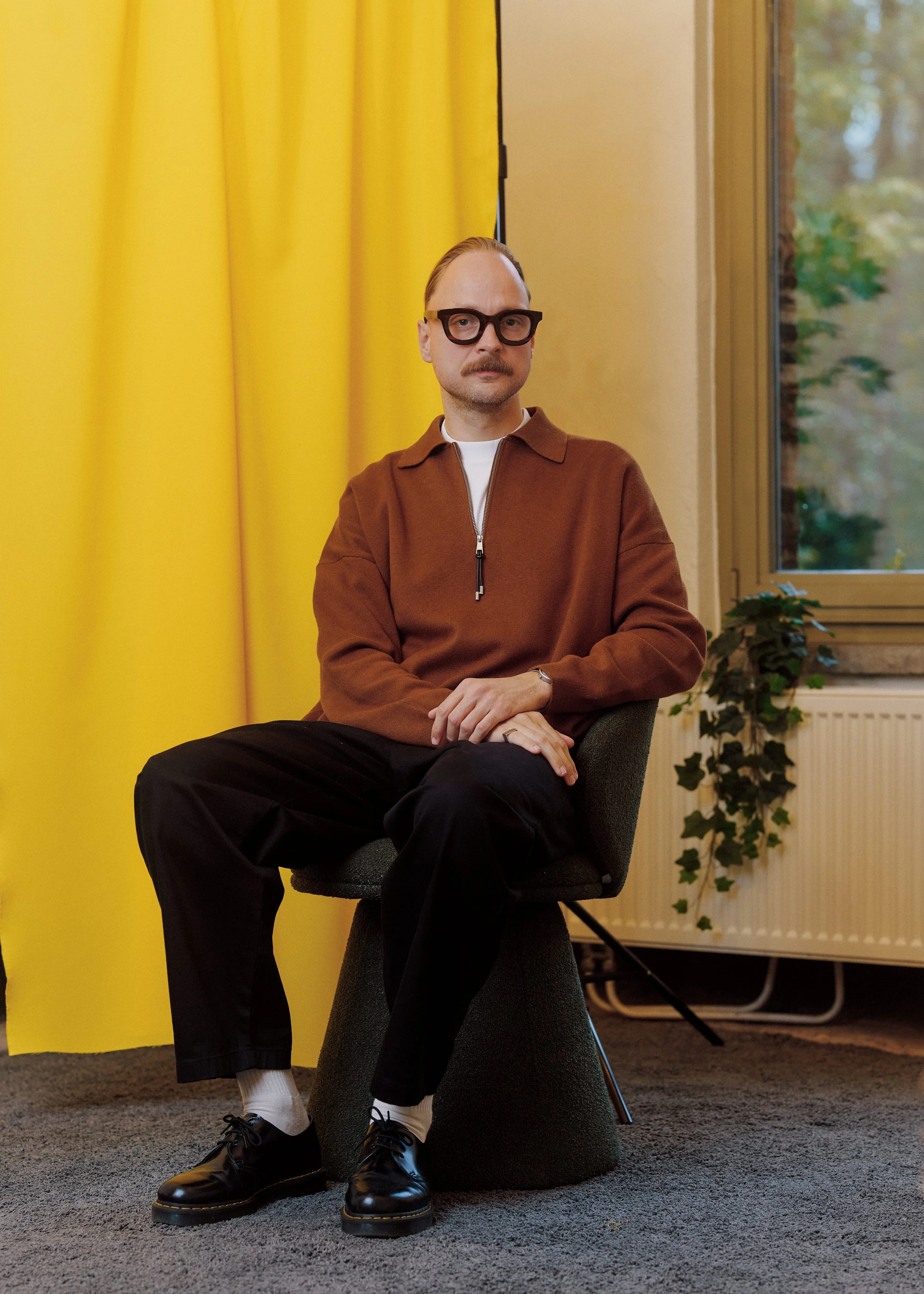 A man with glasses, a mustache, and a receding hairline sitting on a gray ottoman in front of a bright yellow curtain in a room with light yellow walls and a window showing green trees outside.