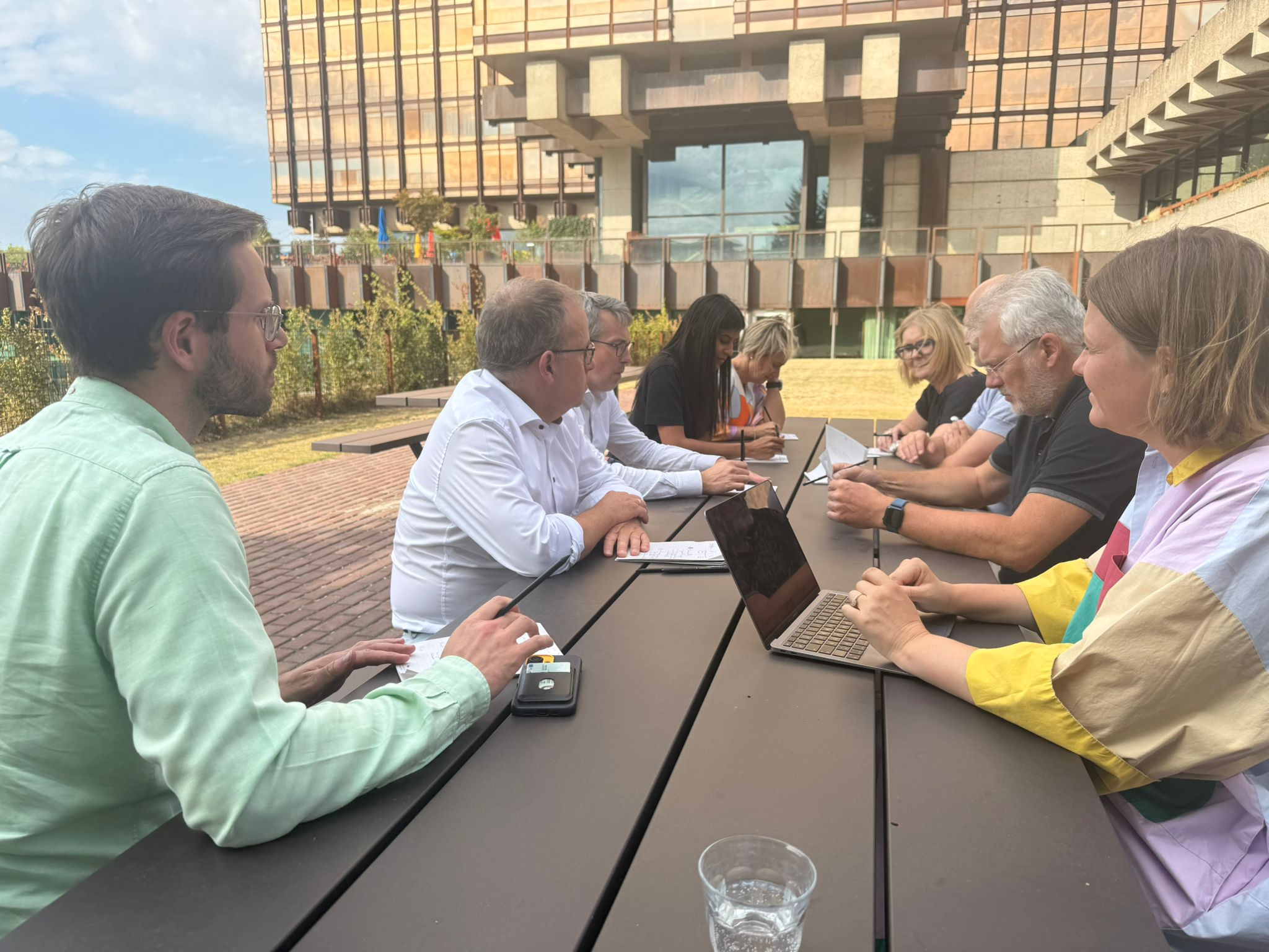 A group of people sitting at a long outdoor table having a meeting or discussion, some taking notes and using laptops, with a modern building in the background.