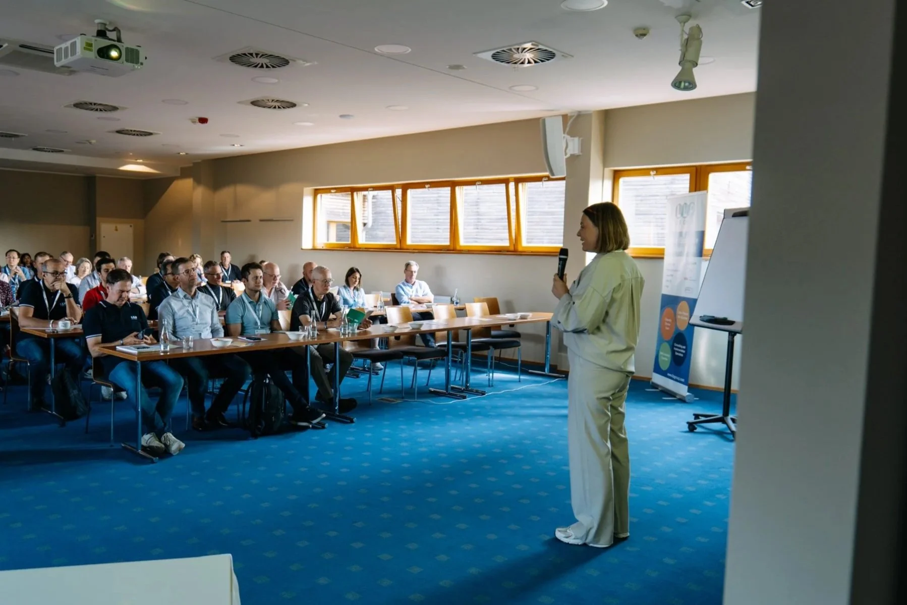 A woman in white clothes holding a microphone presents to an audience in a conference room with large windows, blue carpet, and a whiteboard.