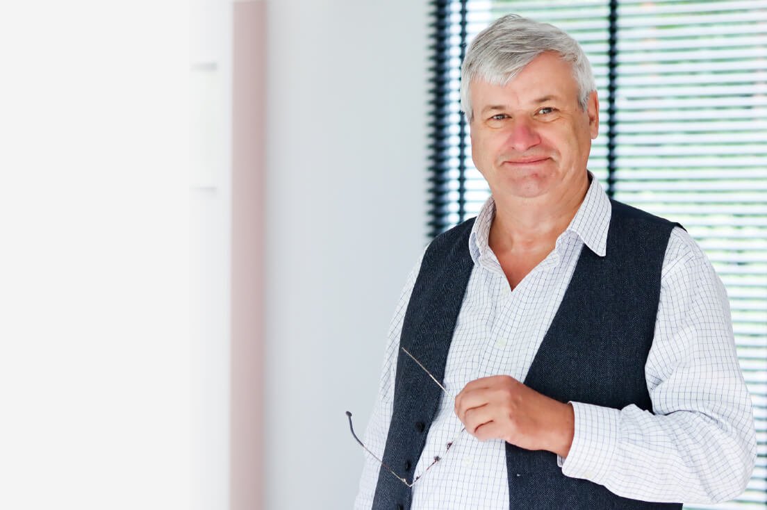 A middle-aged man with gray hair smiles while holding his glasses in an office setting with window blinds in the background.