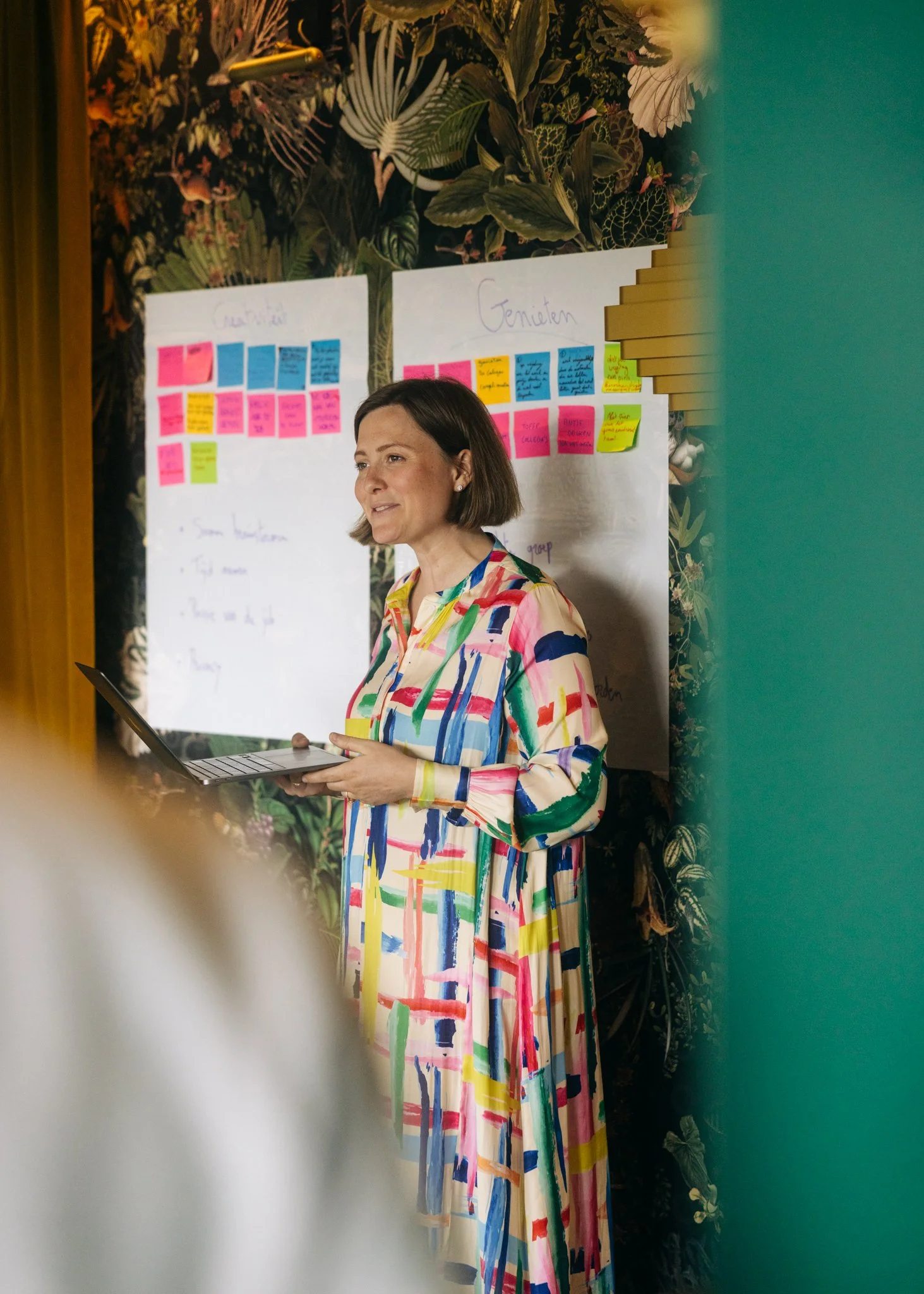 A woman with short brown hair wearing a colorful, abstract-patterned dress is holding a laptop and speaking in front of a whiteboard with sticky notes. The background features tropical-themed wallpaper with large leaves and plants.