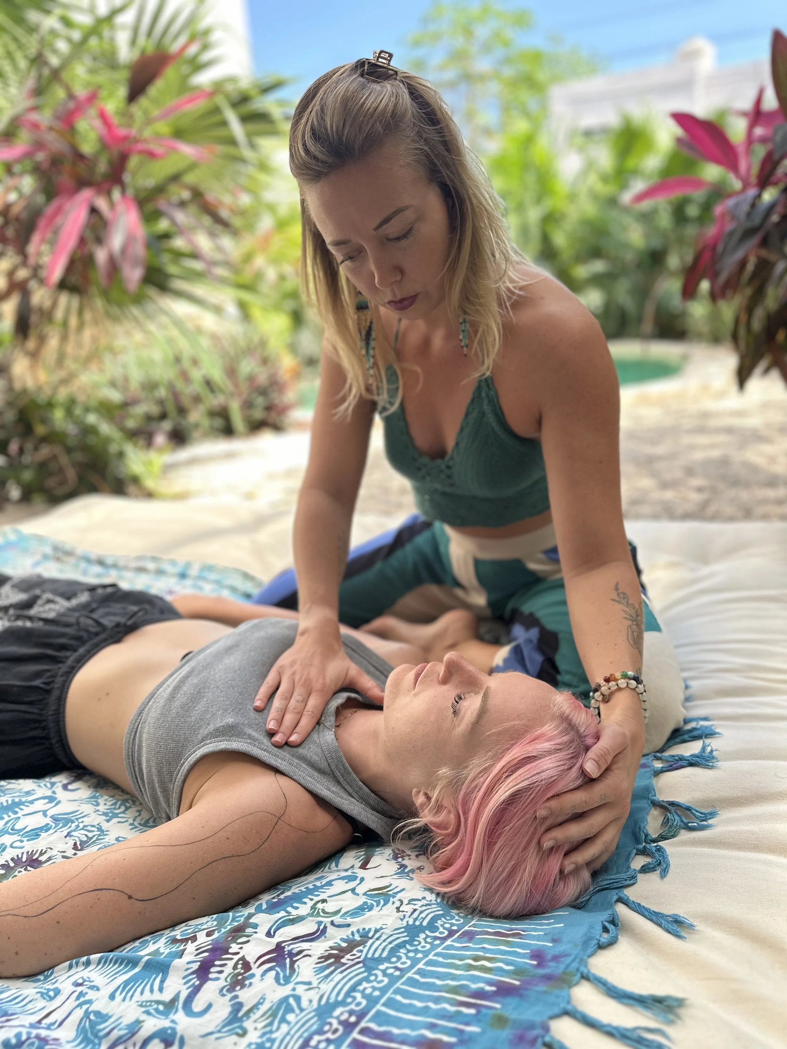 A woman with blonde hair giving a bodywork session to another woman lying on a colorful blanket outdoors with lush green plants in the background.