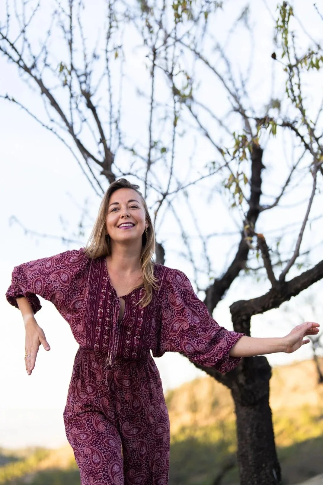 A woman wearing a patterned maroon dress standing outdoors with her left arm extended and a smile on her face, in front of a tree with bare branches.