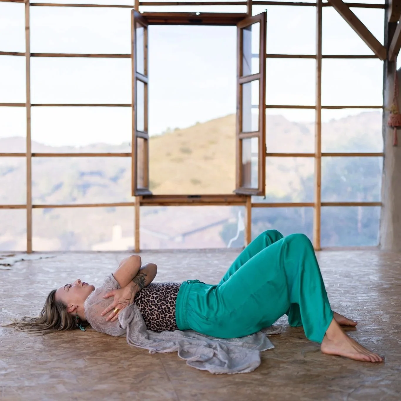 Woman lying on her back inside a room with large open windows showing mountains in the background, arms crossed over her chest, wearing turquoise pants and a leopard-print top.