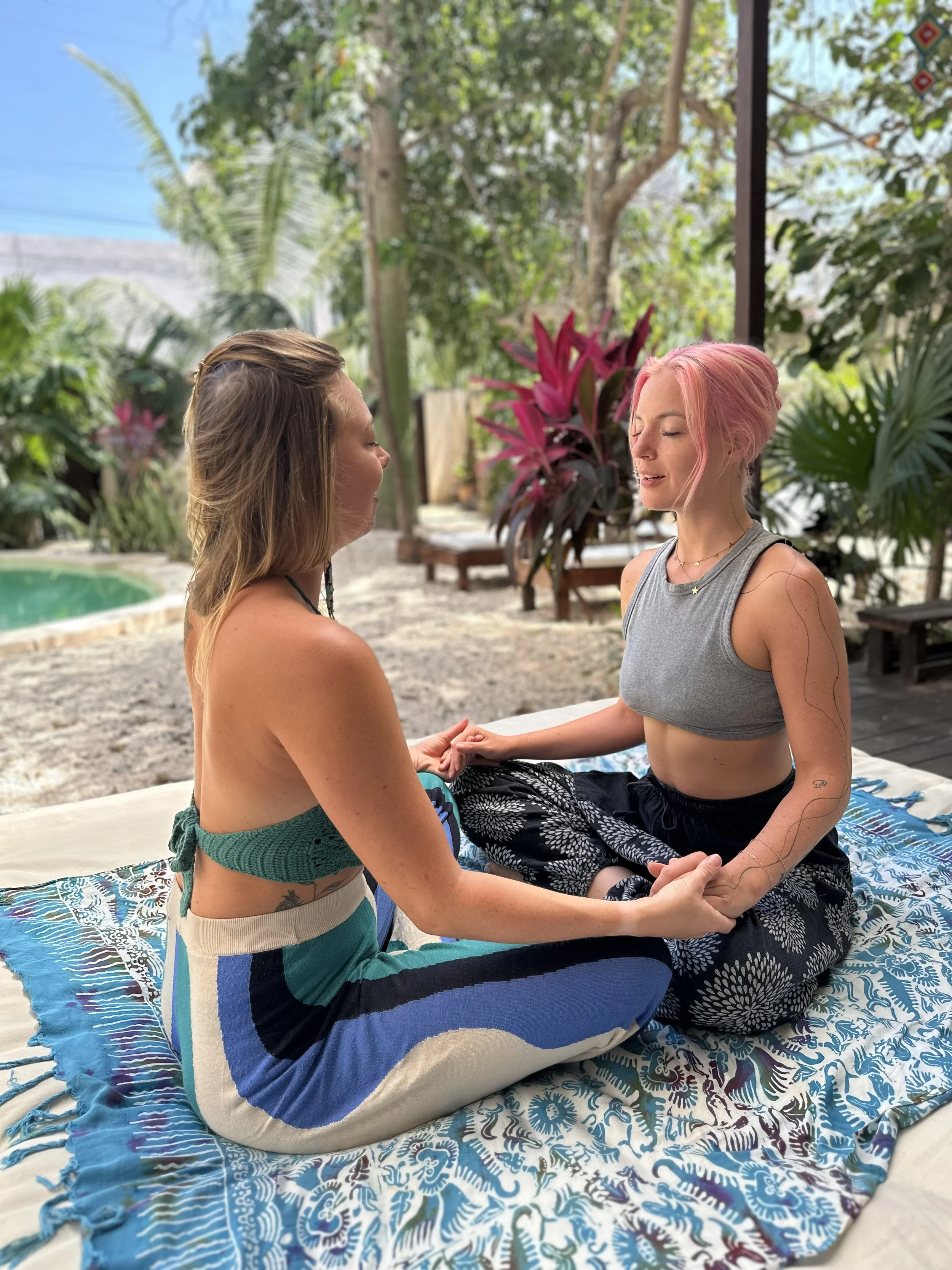 Two women sitting cross-legged on a patterned blanket outdoors, holding hands and facing each other with eyes closed, surrounded by lush tropical plants and trees.