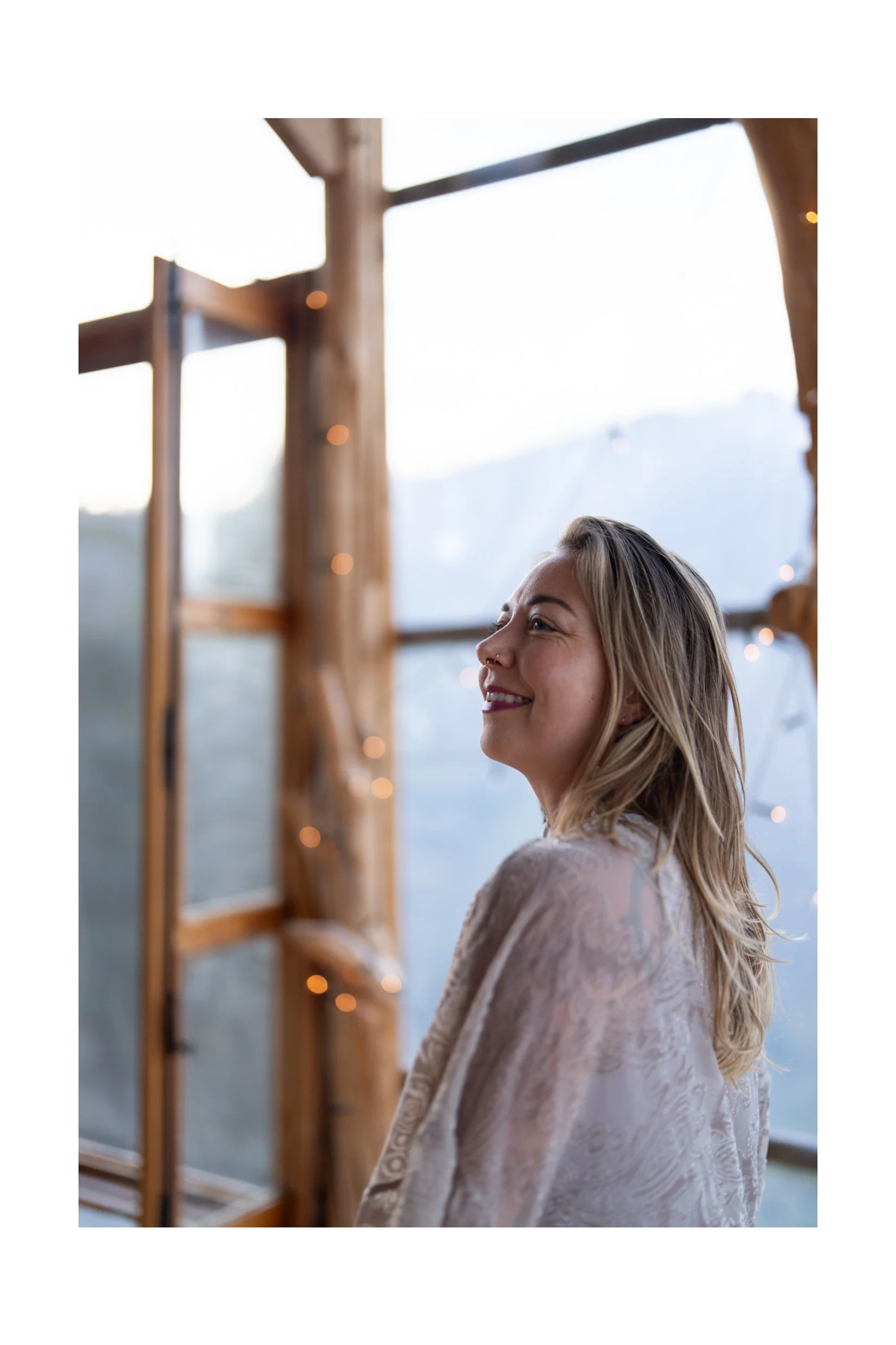 A woman with blonde hair smiling indoors near a window with mountain scenery in the background, decorated with string lights.