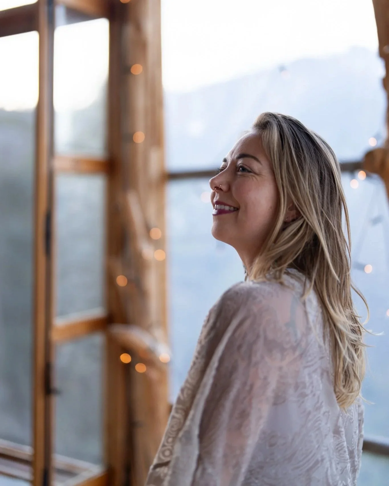 A woman with blonde hair smiling and looking to the right inside a wooden structure decorated with small string lights, with a blurred mountain landscape in the background.