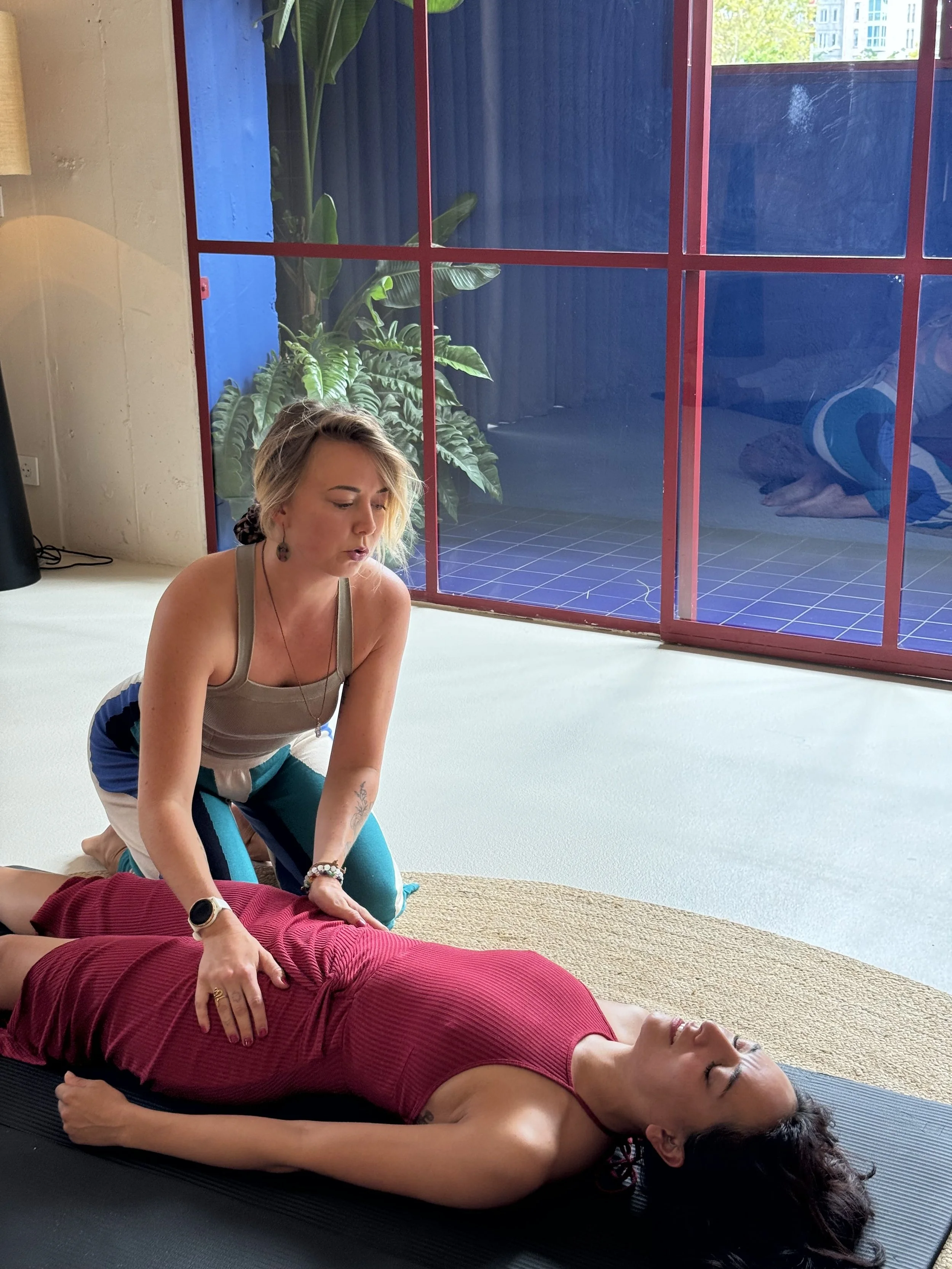 A woman in a red dress receiving an energy healing session from a practitioner in a yoga studio with large windows and plants.