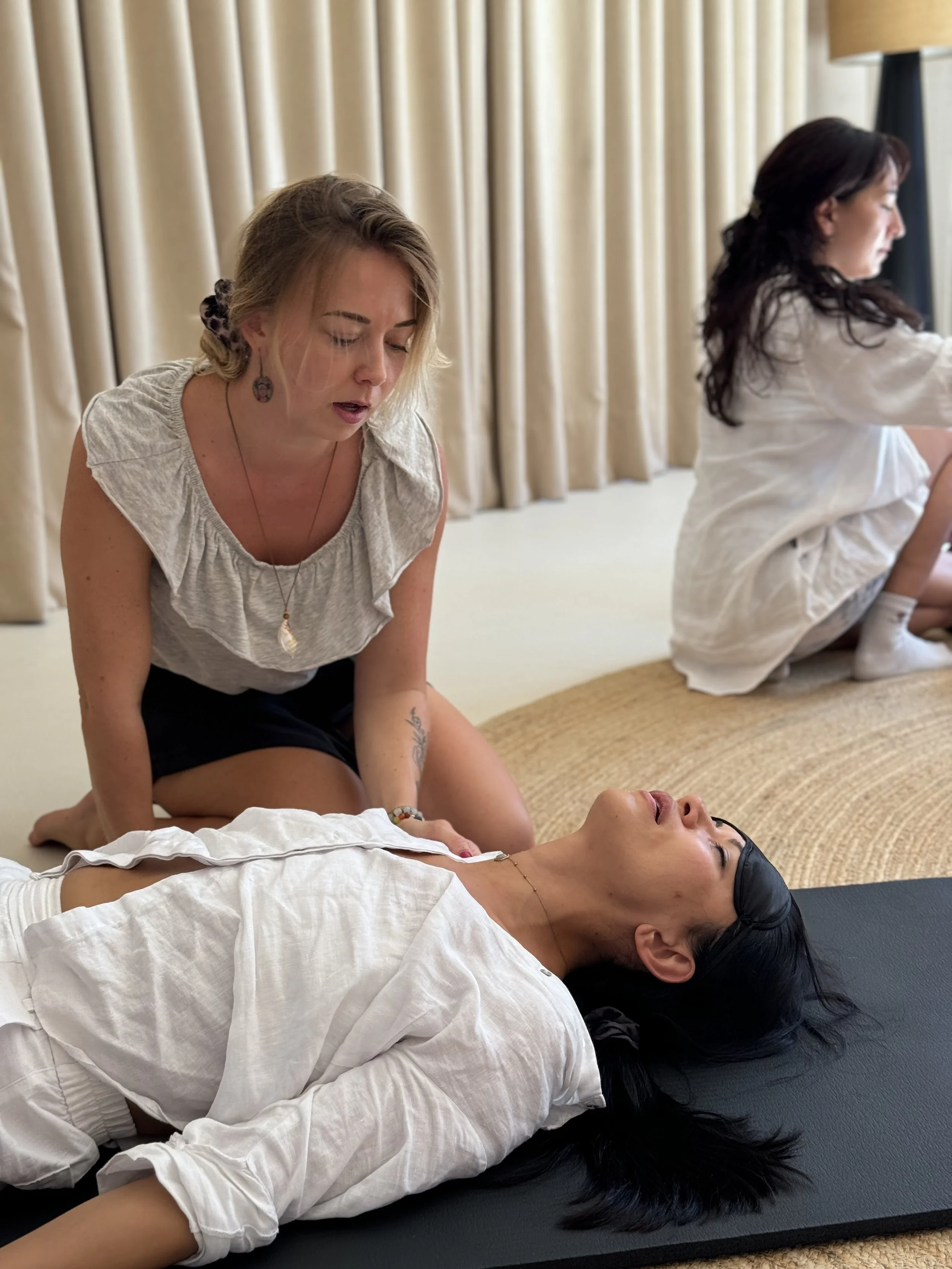 A woman is lying down on a yoga mat with her eyes closed, while another woman is kneeling beside her, appearing to perform a healing or massage technique. A third woman is sitting on the floor in the background, near beige curtains.
