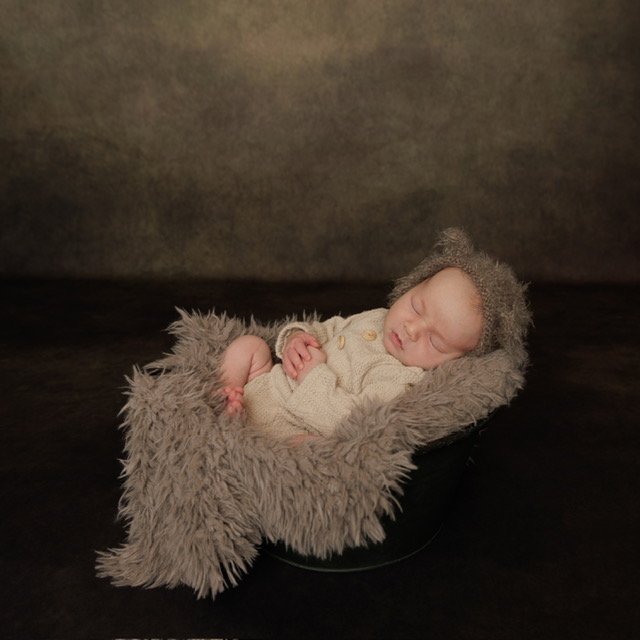 A newborn baby sleeping in a cozy basket with a fuzzy blanket and a knit hat, against a dark background. Professional photography at Studio 5 by JK Photography, Carlisle UK