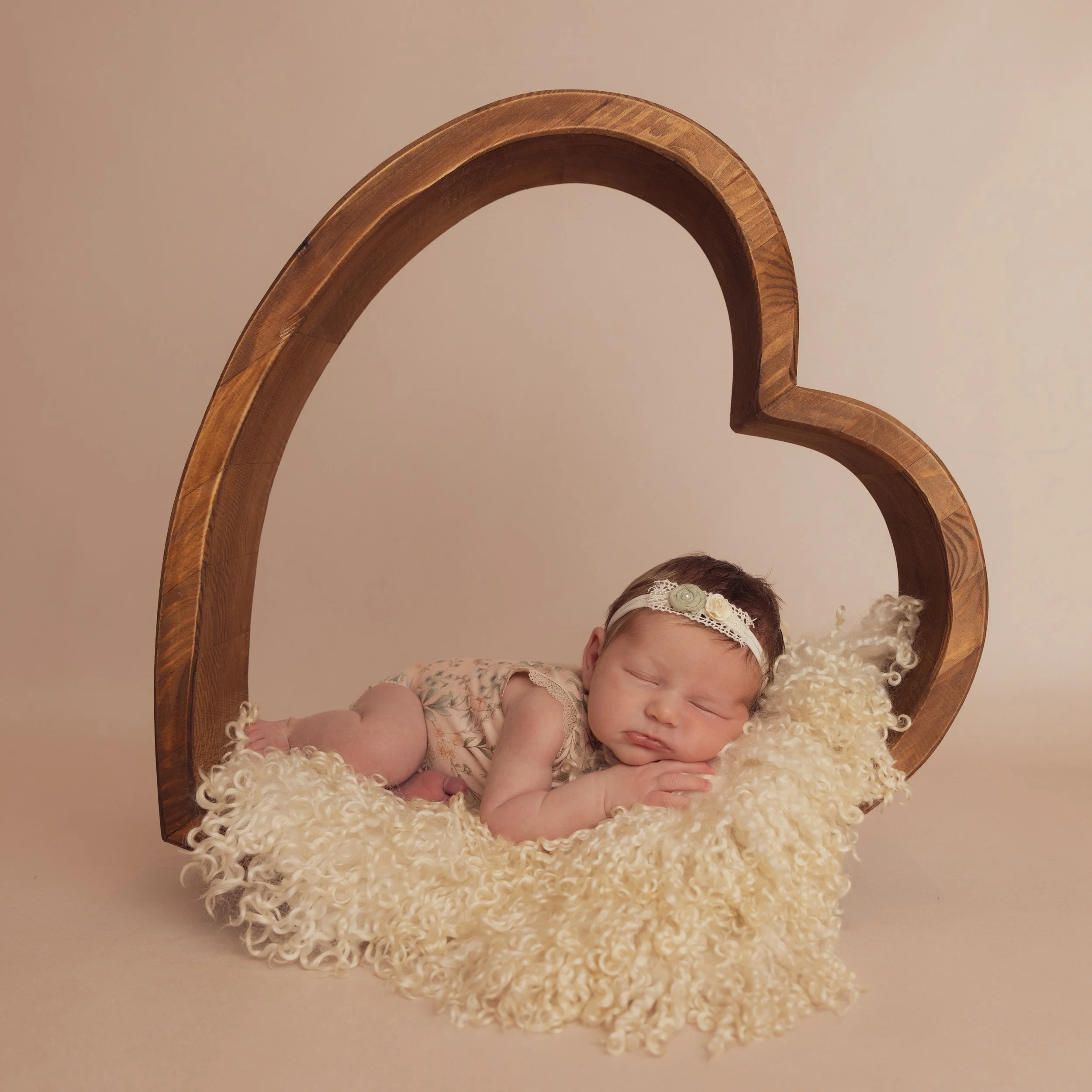 A sleeping baby girl lying on a fluffy cream blanket inside a large wooden heart-shaped frame. Professional photography at Studio 5 by JK Photography, Carlisle, Cumbria.