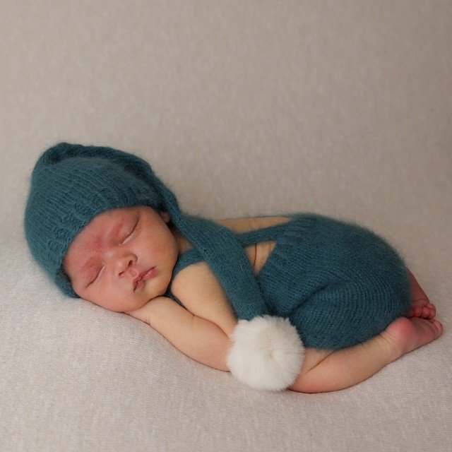 A newborn baby sleeping, dressed in a teal knit hat with a pom-pom, teal sweater, and matching pants, curled up on a light-colored surface. Professional photography at Studio 5 by JK Photography, Carlisle, Cumbria.