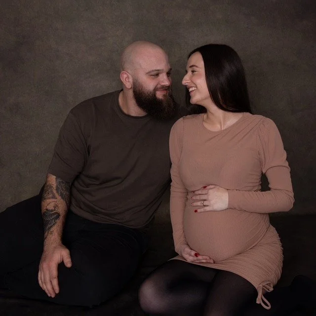 A pregnant woman in a beige dress sits next to a man with a beard and tattoos on his arm. They are looking at each other and smiling, with a neutral dark background behind them.