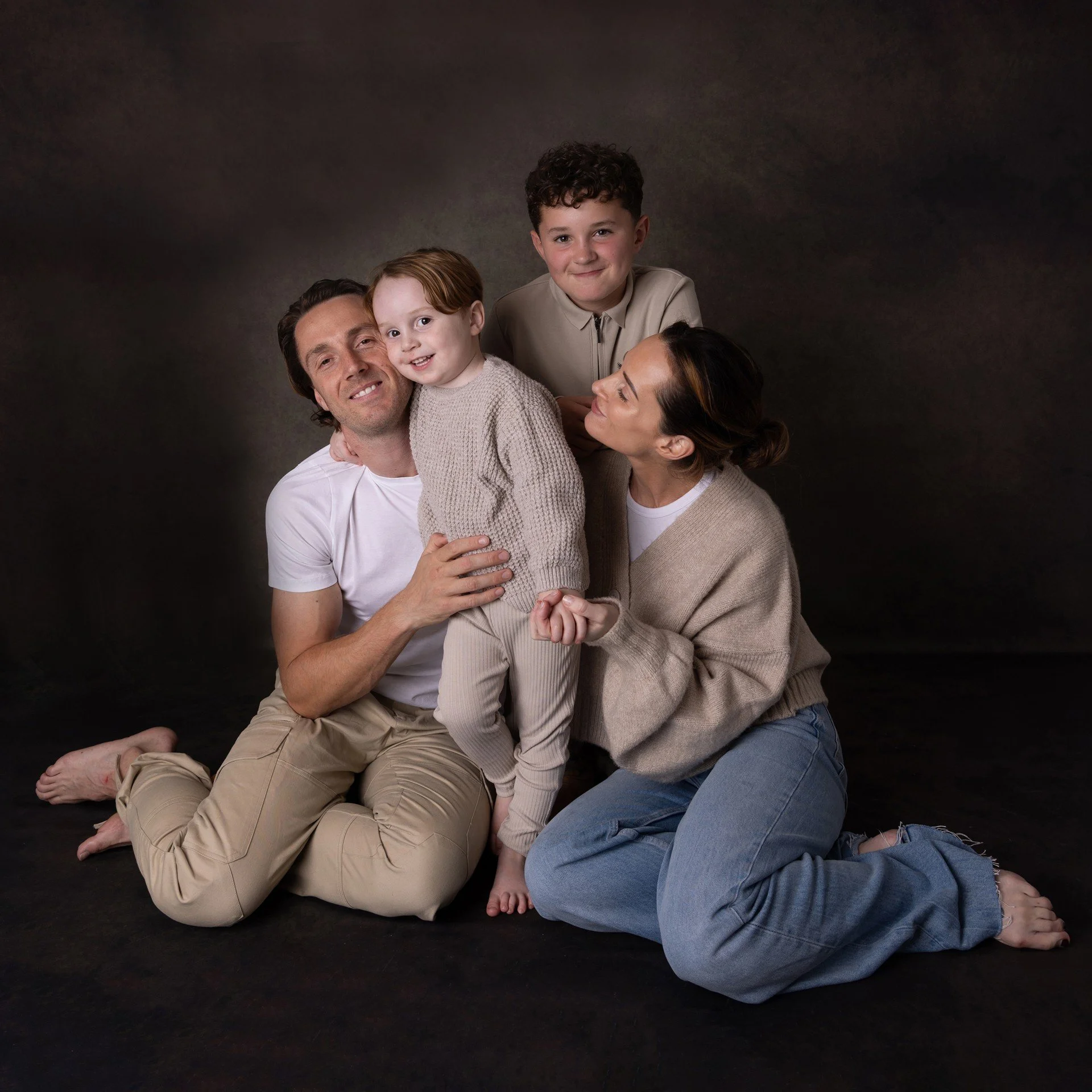 A family of four, including two adults and two children, posing together on a dark studio background. The adults are kneeling while the children are standing, with everyone smiling and close together.