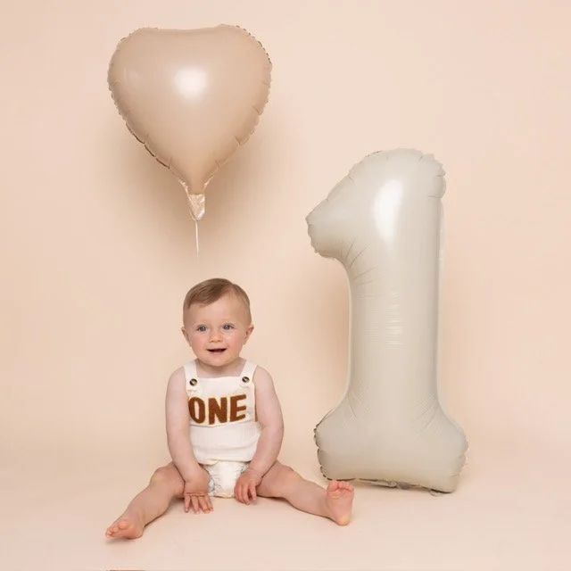 A baby sitting on the floor with two balloons, a heart-shaped balloon and a number one-shaped balloon, for a first birthday celebration.