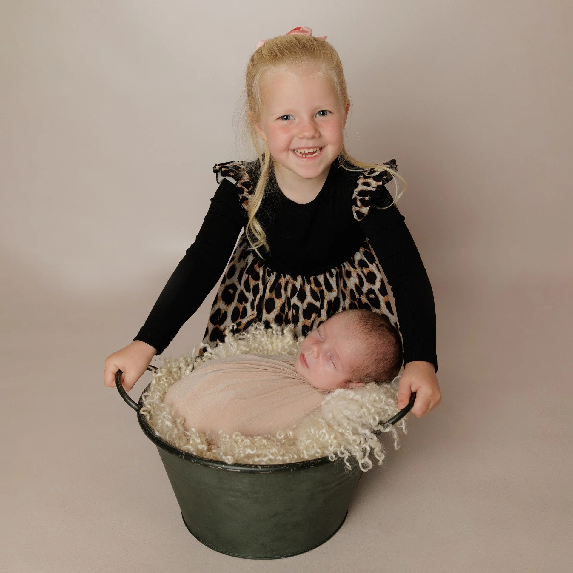 A young girl with blonde hair wearing a black and leopard print dress, is smiling and holding a black bucket with a new born baby wrapped in a beige cloth and surrounded by fluffy white material. Professional photography by JK Photography.