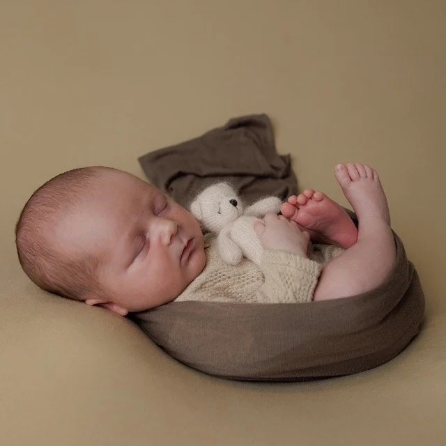 A sleeping baby wrapped in a brown blanket, holding a small stuffed animal, lying on a beige surface. Professional photography at Studio 5 by JK Photography, Carlisle.