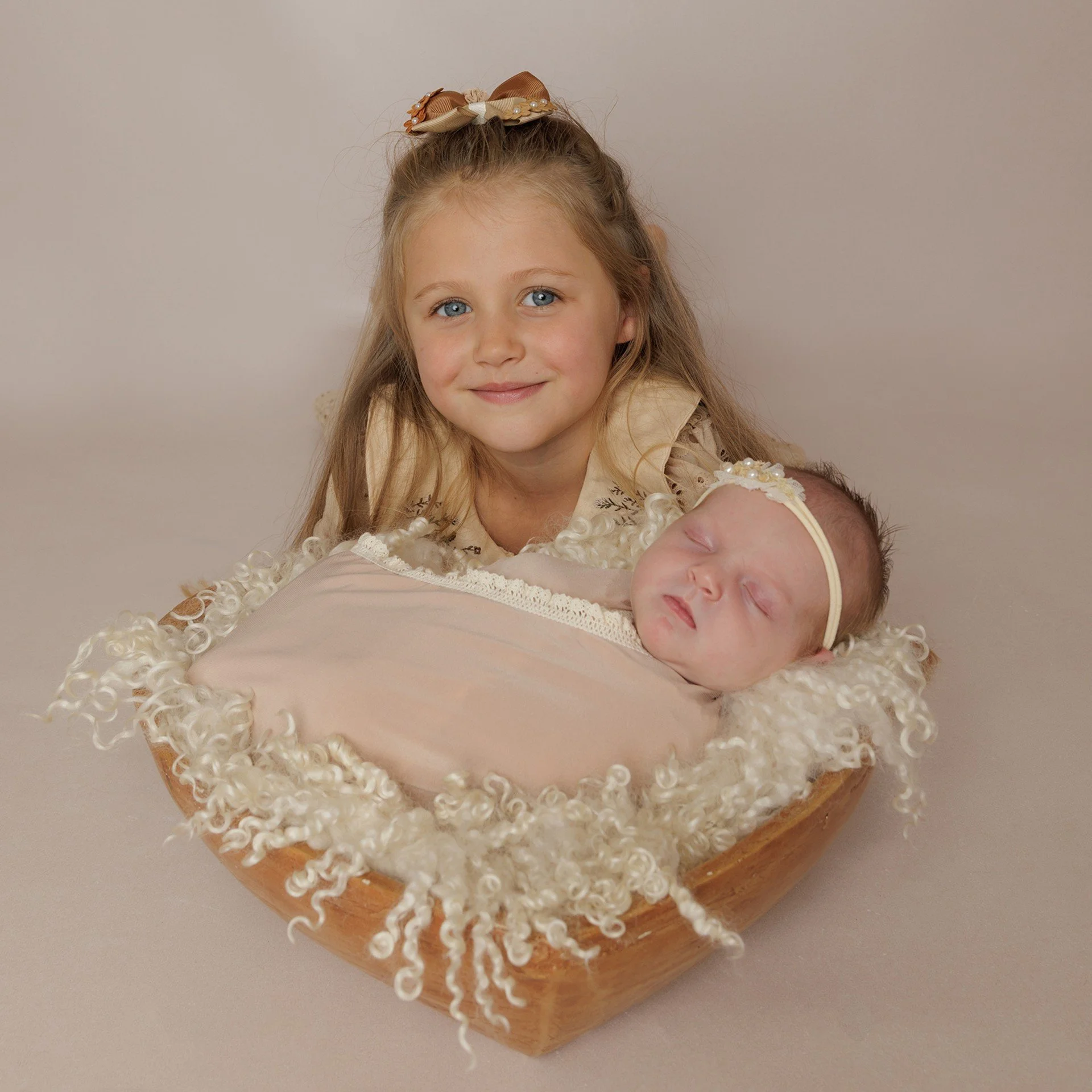 A young girl with blonde hair and a bow in her hair holds a sleeping baby wrapped in a pink blanket in a wooden bowl lined with curly white fabric. Photography in the Gallery at JK Photography Carlisle Cumbria. 