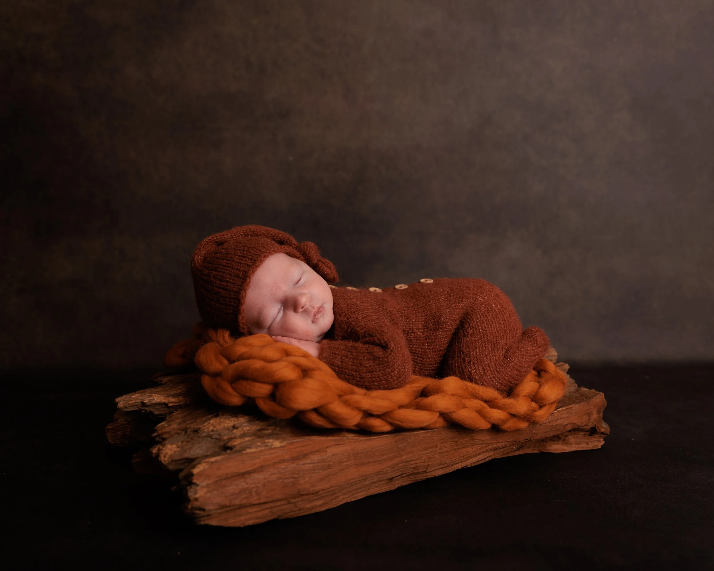 A sleeping newborn baby dressed in a brown knitted outfit and hat, lying on a woven orange blanket on a rustic wooden slab against a dark background.