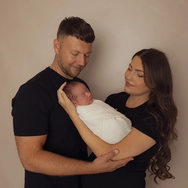 A man and woman holding a sleeping newborn baby wrapped in a white blanket, with the man smiling and looking at the baby and the woman looking at the baby affectionately. Photographed at Studio 5 at JK Photography Carlisle.