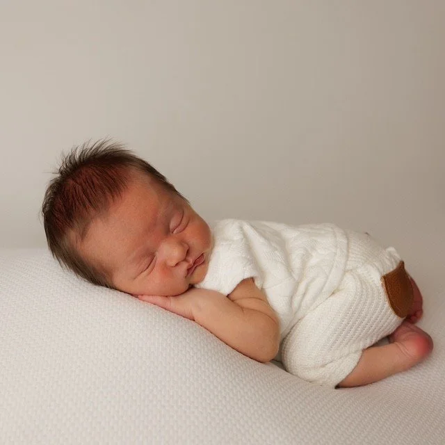 A sleeping baby lying on a white surface, wearing a white shirt and white pants.