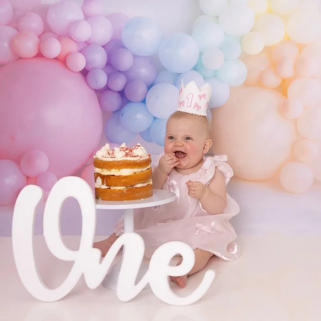 Baby girl celebrating her first birthday sitting next to a cake with colorful balloons in the background, wearing a birthday crown and a pink dress.