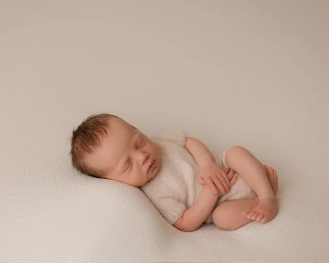 Example of new born photography in the Gallery at JK Photography Carlisle Cumbria. Picture of a sleeping infant baby lying on a white textured surface, wearing a light-colored outfit.