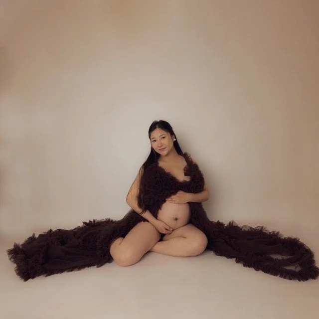 A pregnant woman sitting on the floor in front of a plain wall, wearing a dark, fluffy garment and smiling at the camera. Photographed at Studio 5 by JK Photography, Carlisle, Cumbria.