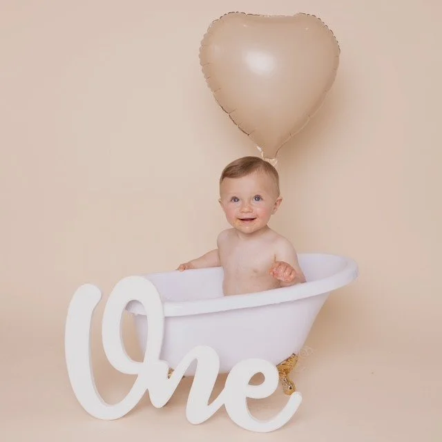 A smiling baby sitting in a small white bathtub with a gold balloon shaped like a heart floating above. The baby is next to a large white decorative sign that spells 'One'.