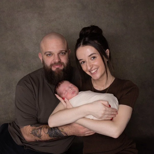 A happy family portrait featuring a bearded man and a smiling woman holding a newborn baby against a plain dark background.
