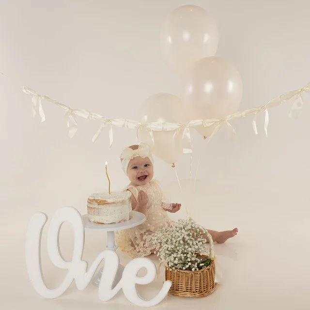Baby celebrating first birthday with a cake, balloons, and a "One" sign in a white studio setting.
